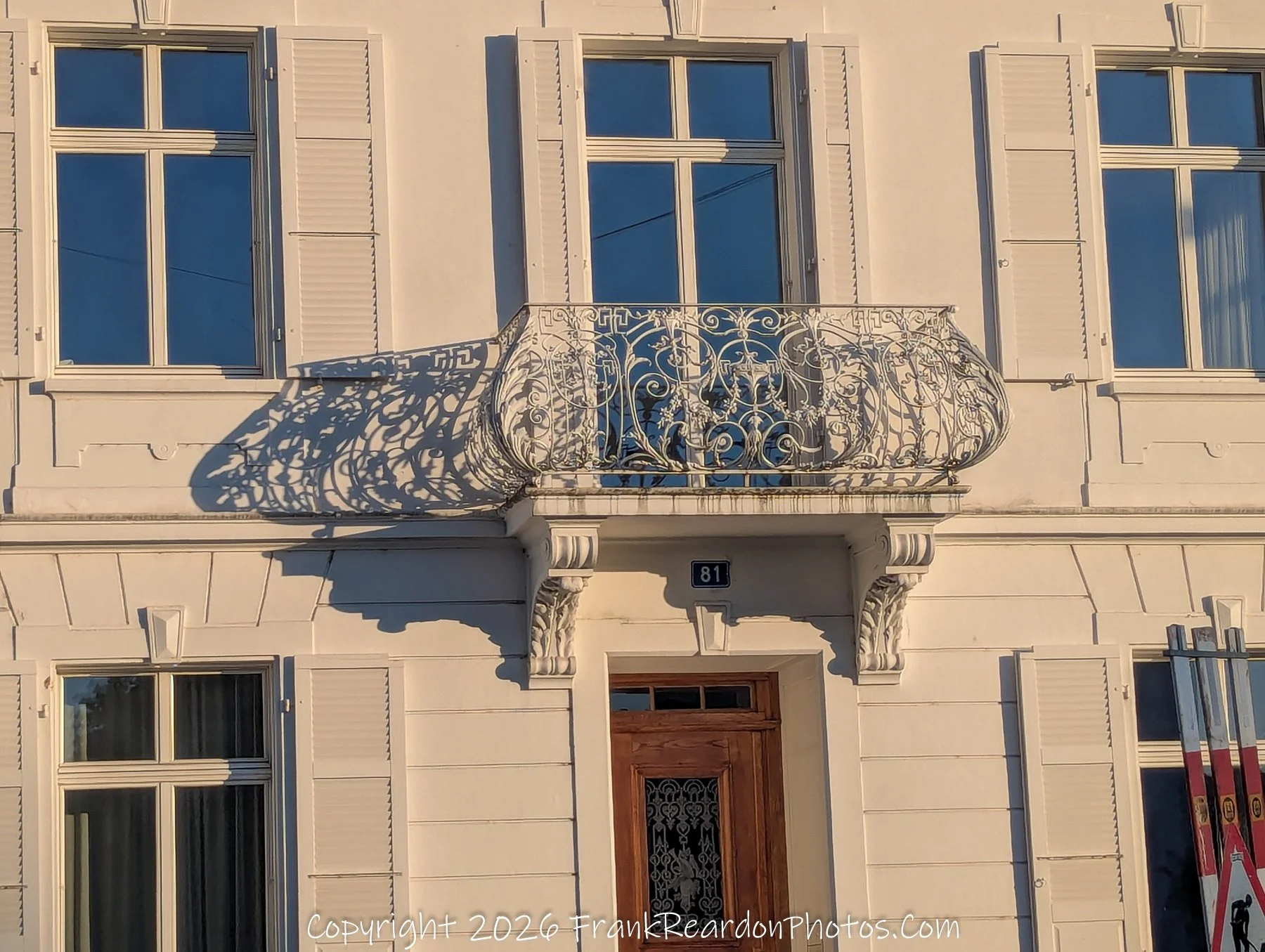 "Ornate Balcony Shadow" A close-up of a white building façade with a detailed, wrought-iron balcony railing that casts a complex, intricate shadow onto the wall behind it.