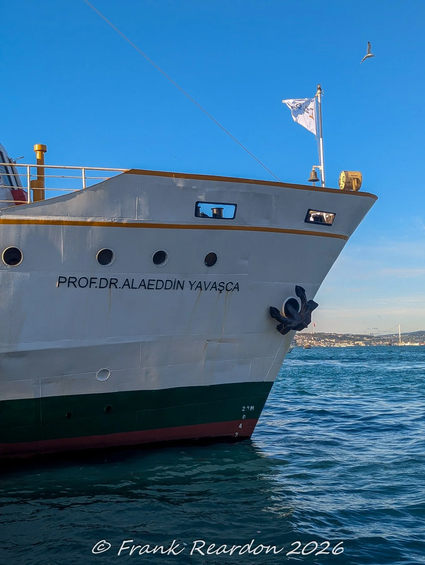 The bow of the municipal passenger ferry Prof. Dr. Alaeddin Yavaşça cutting through the blue waters of the Bosphorus, complete with an anchor and a fluttering flag.