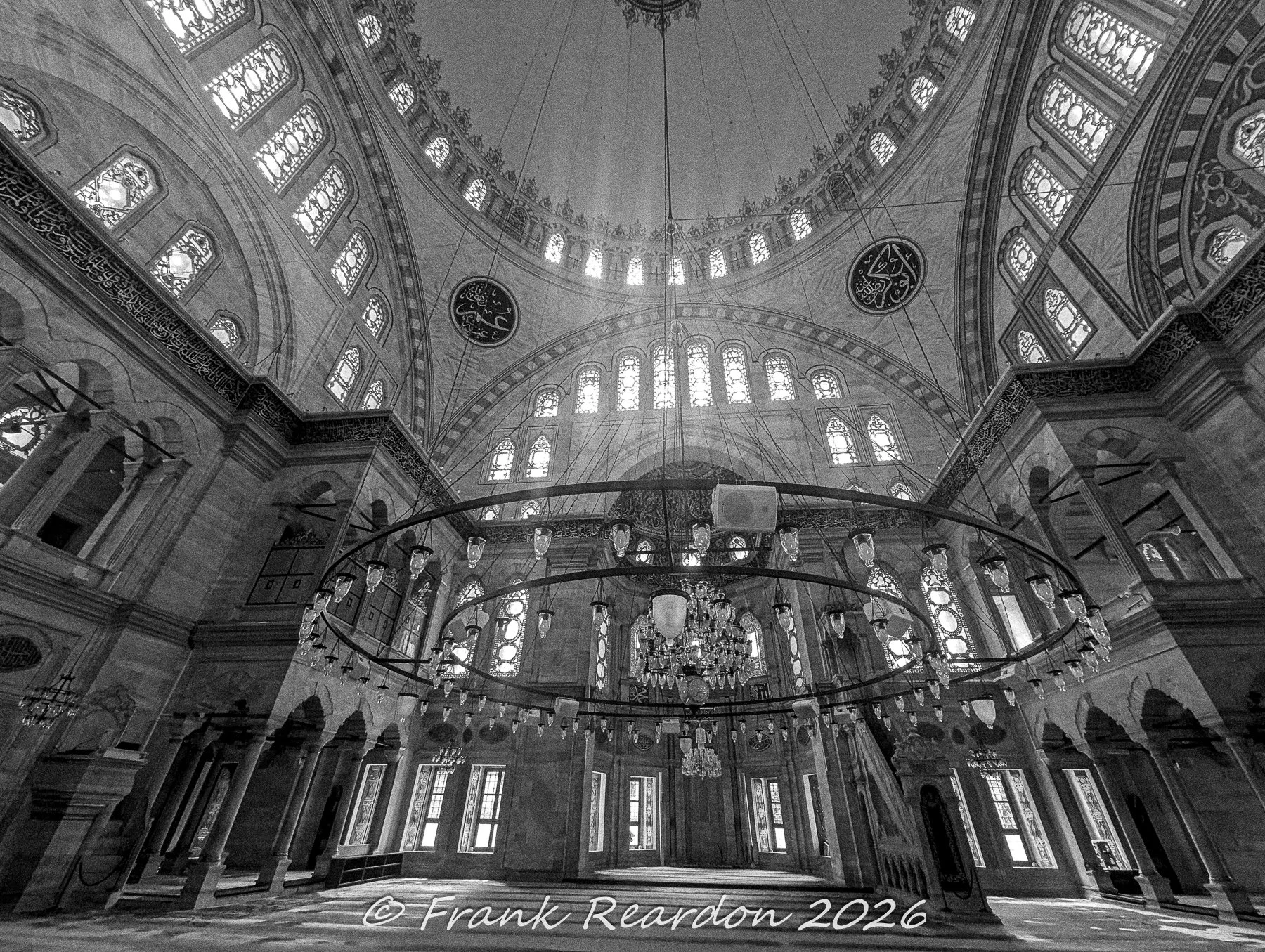 A dramatic black-and-white perspective highlighting the grand scale, stone arches, and detailed ring chandelier inside the Nuruosmaniye Mosque.