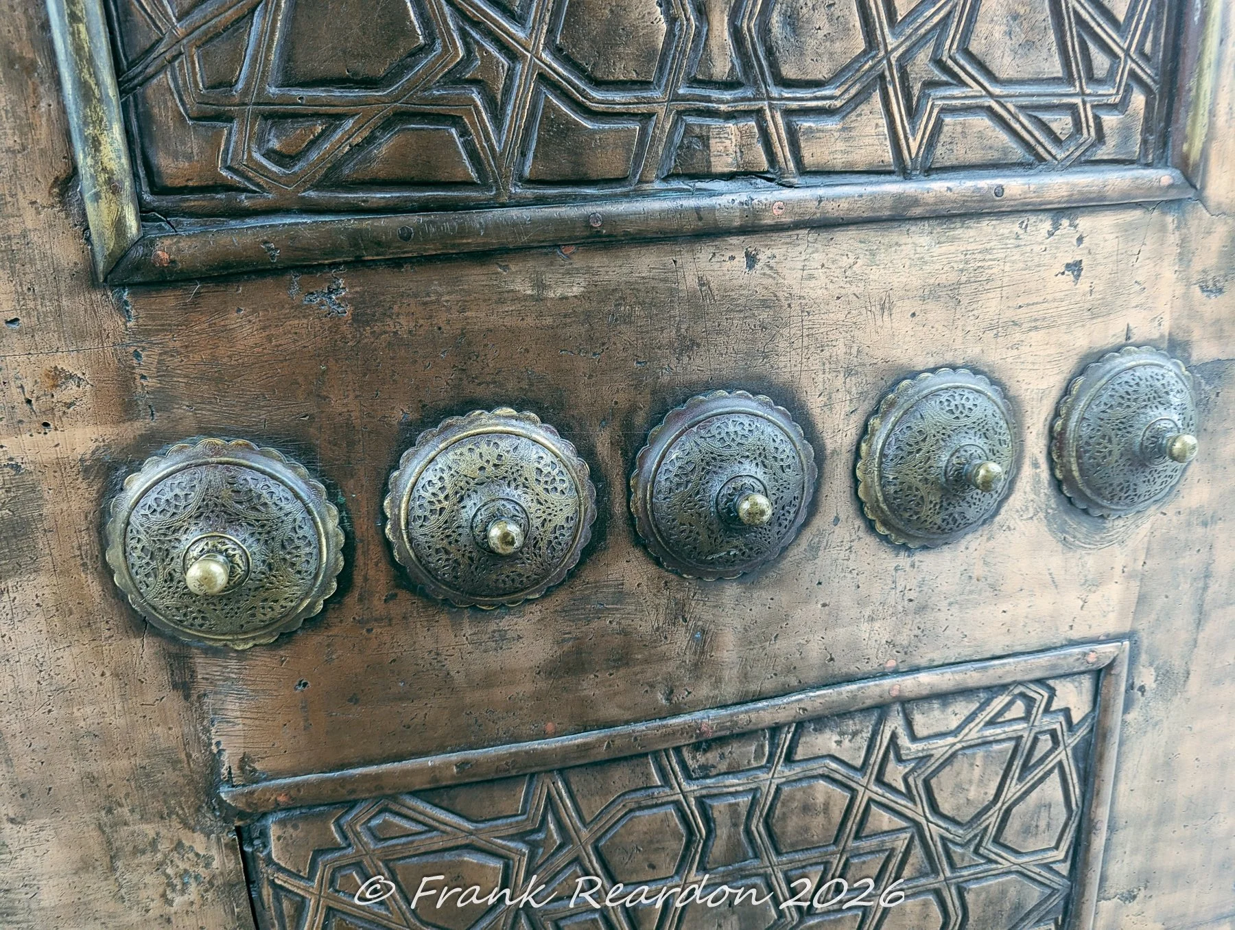 A detailed shot of traditional geometric wood carving and decorative brass doorknobs on a historic door within the Blue Mosque complex.