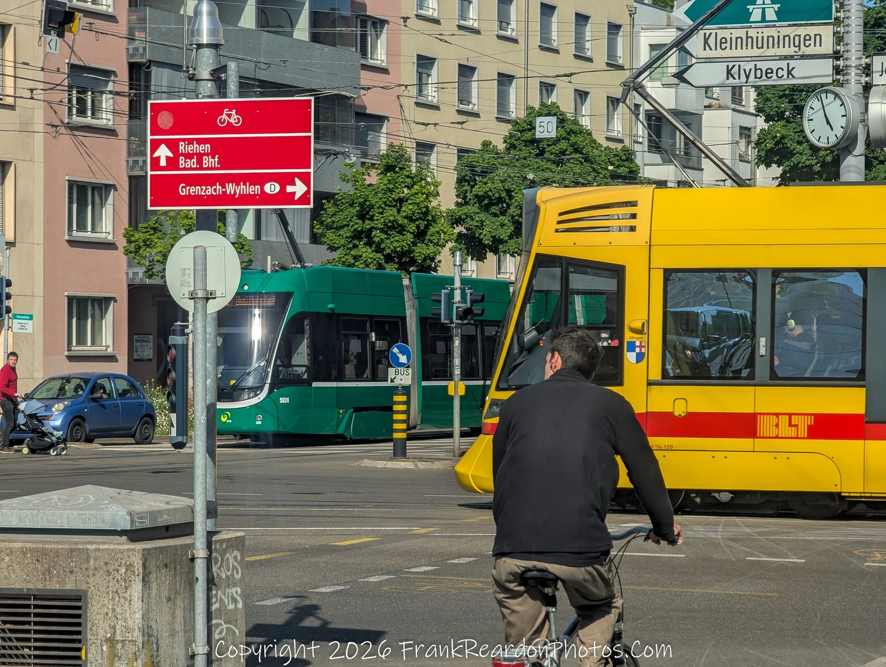 "Urban Intersection: Trams, Bikes, and Signs"
A busy city street corner featuring a cyclist, a yellow public transit tram, a green tram, and directional signage for local destinations.