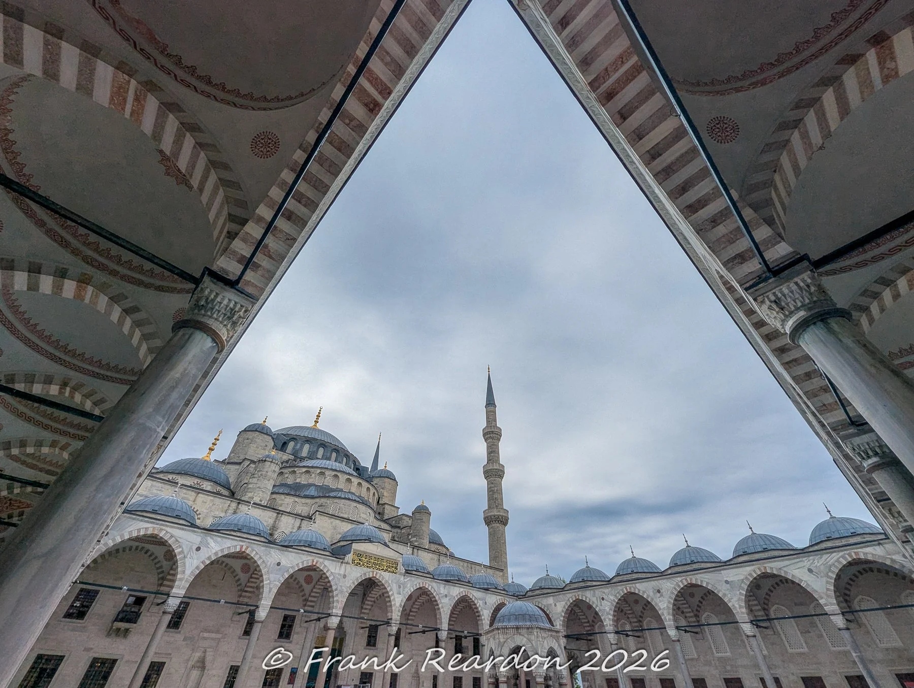 A geometric perspective from beneath the arcades of the Sultan Ahmed Mosque (Blue Mosque) framing the central courtyard and a towering minaret against the sky.