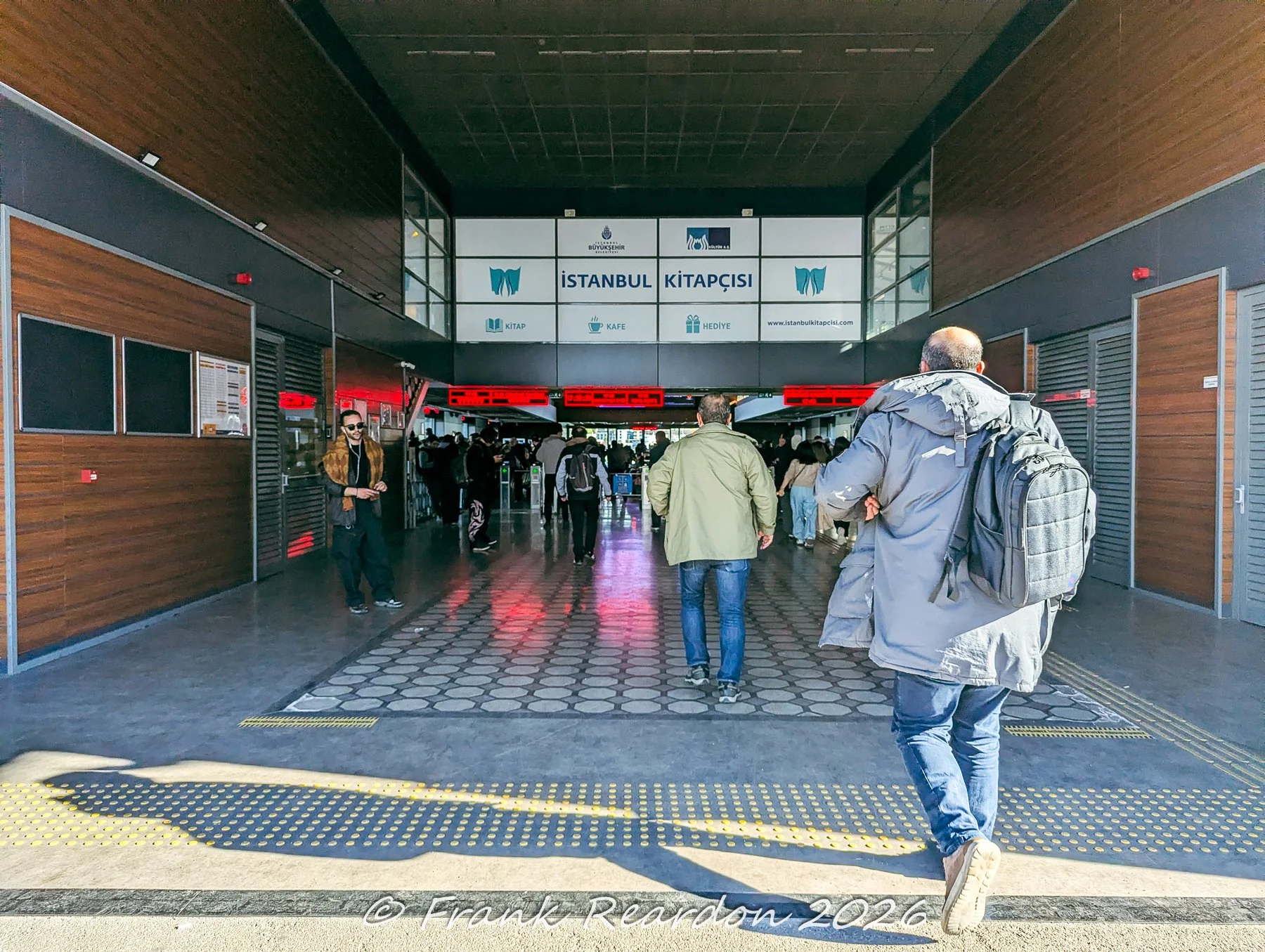 People walking through a modern transit concourse and bookstore corridor featuring an Istanbul Kitapçısı branch with sleek wooden paneling and signage.