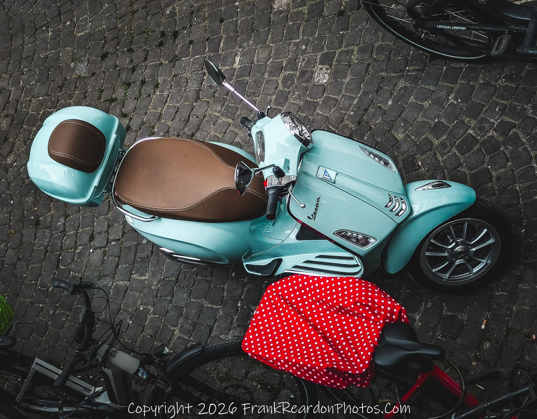 "A Scooter on Cobblestone" A high-angle shot of a light blue Vespa-style scooter with a brown leather seat, parked on traditional dark cobblestone pavement alongside a bicycle draped with a red polka-dot cloth.