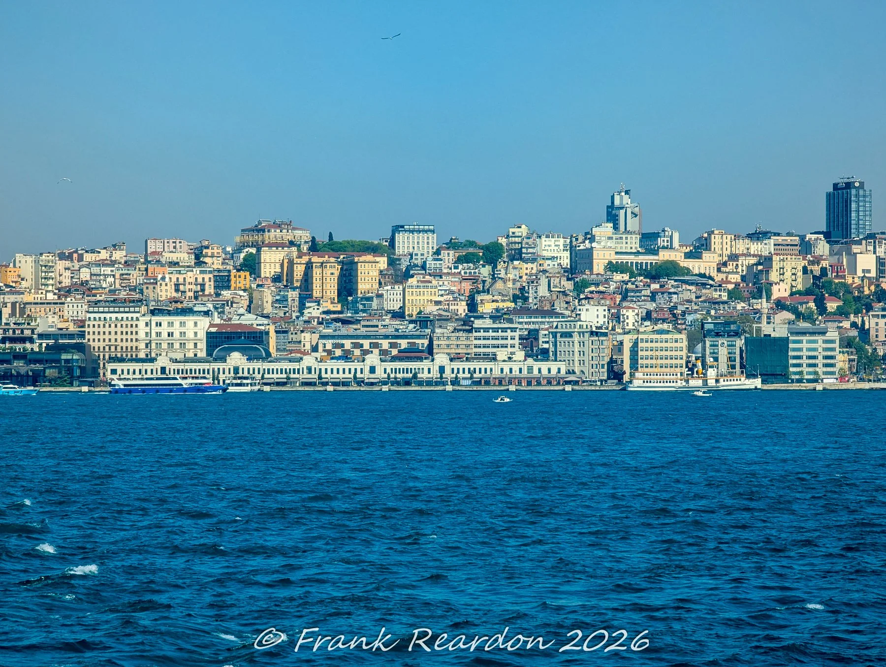 A wide-angle view of the bustling Karaköy coastline, featuring the ferry terminal, waterfront promenades, and the dense layers of Istanbul's urban architecture rising up the hillside.