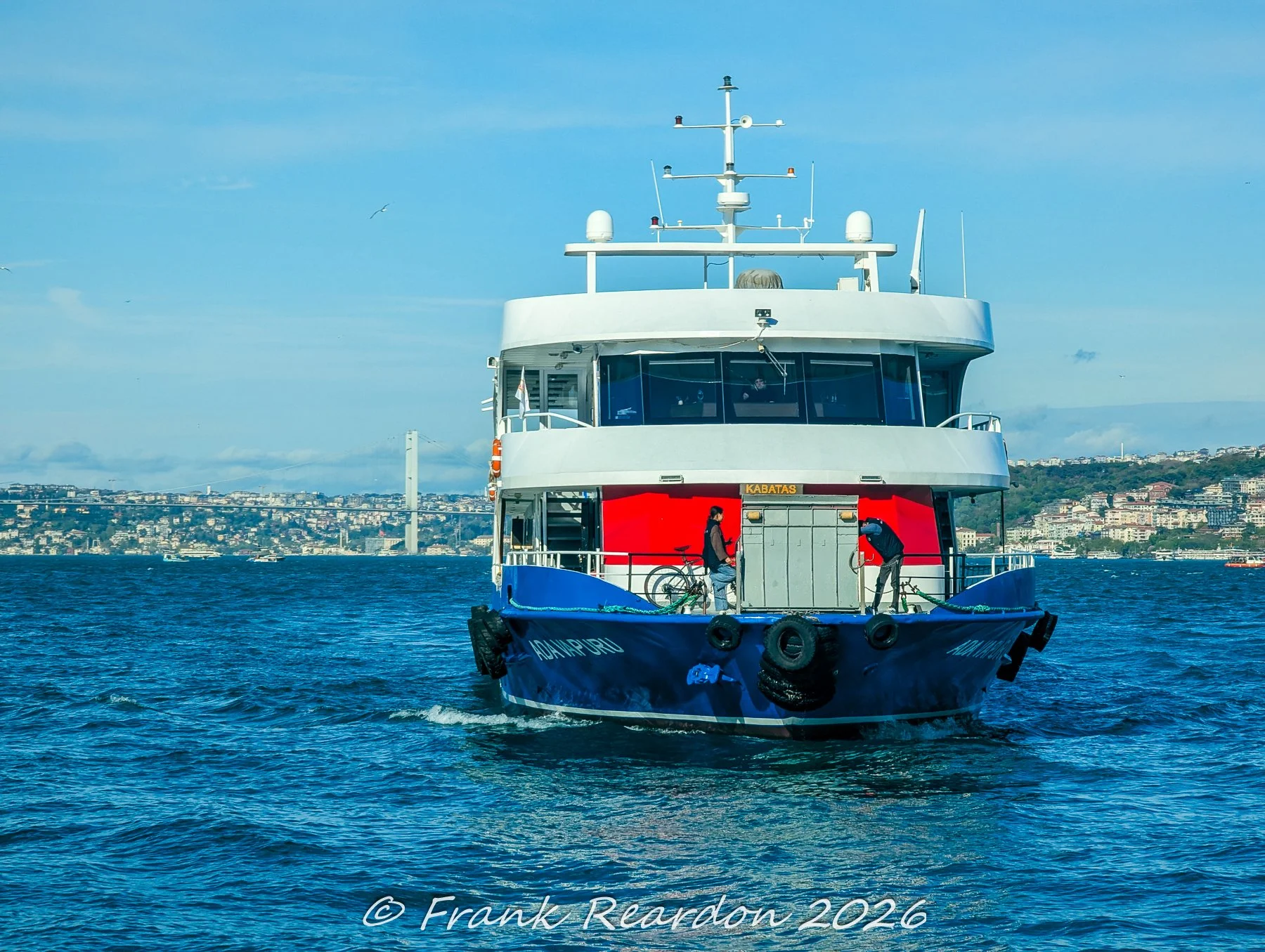 A close-up view of a blue and white urban passenger ferry navigating the waters of the Bosphorus with the city shoreline visible in the background.