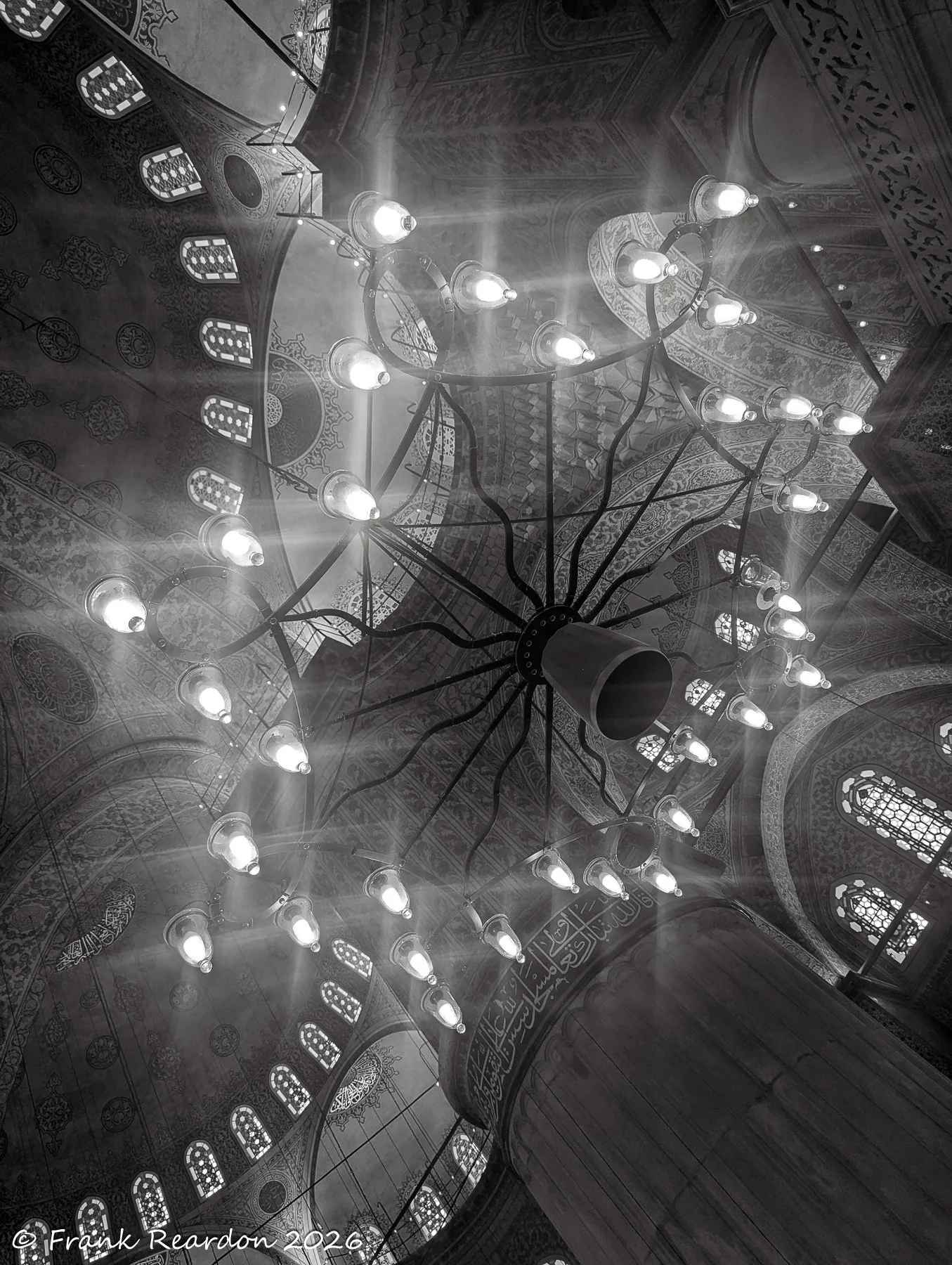 A dramatic low-angle shot looking upward through a glowing chandelier toward the intricate tilework and patterns of the dome.