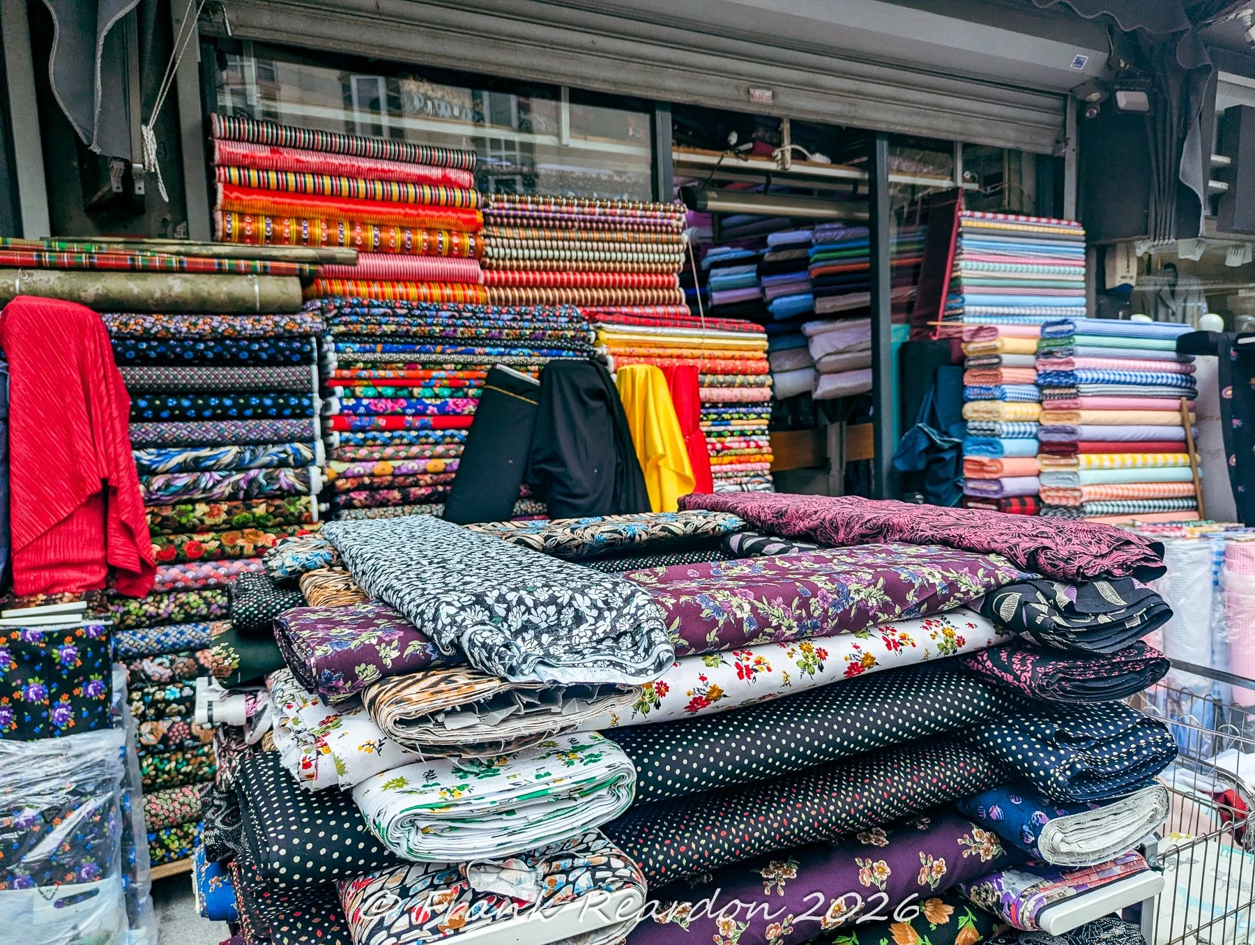A rich display of stacked colorful textiles, patterned rolls of fabric, and folded garments outside a shop in the Grand Bazaar district.