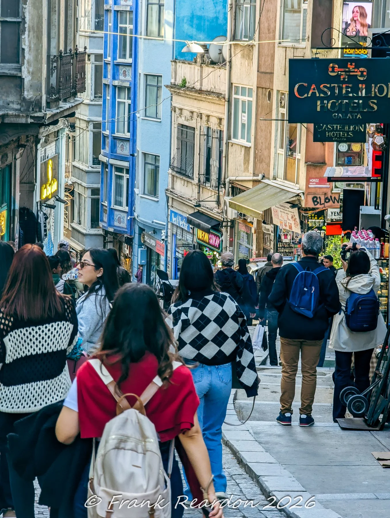 A group of pedestrians walking down a steep, classic hillside street lined with colorful residential buildings and hotel signs.