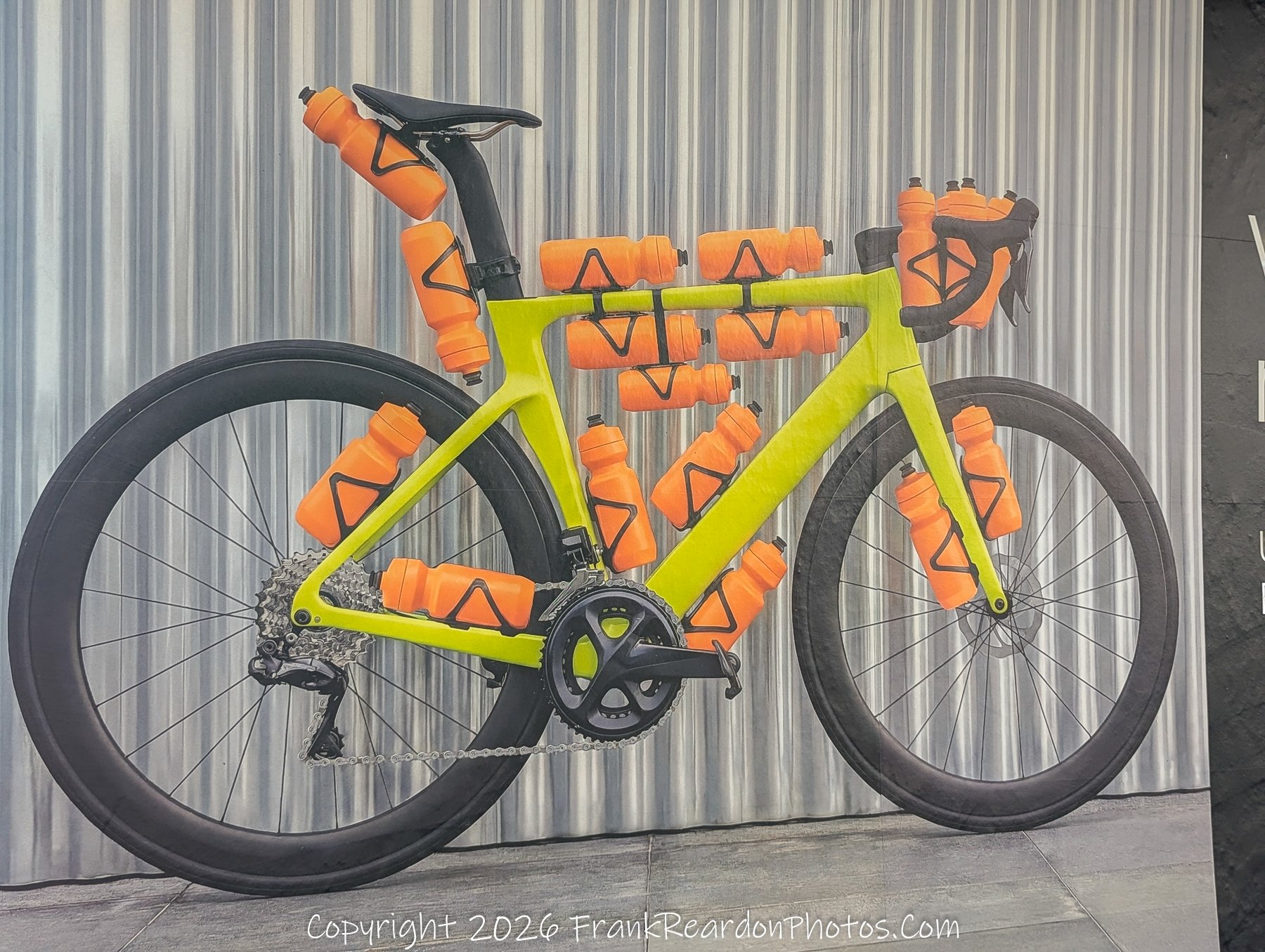 "The Endurance Cyclist’s Display" A promotional or artistic display featuring a lime-green road bike heavily accessorized with numerous bright orange water bottles mounted across its frame, handlebars, and seat post.