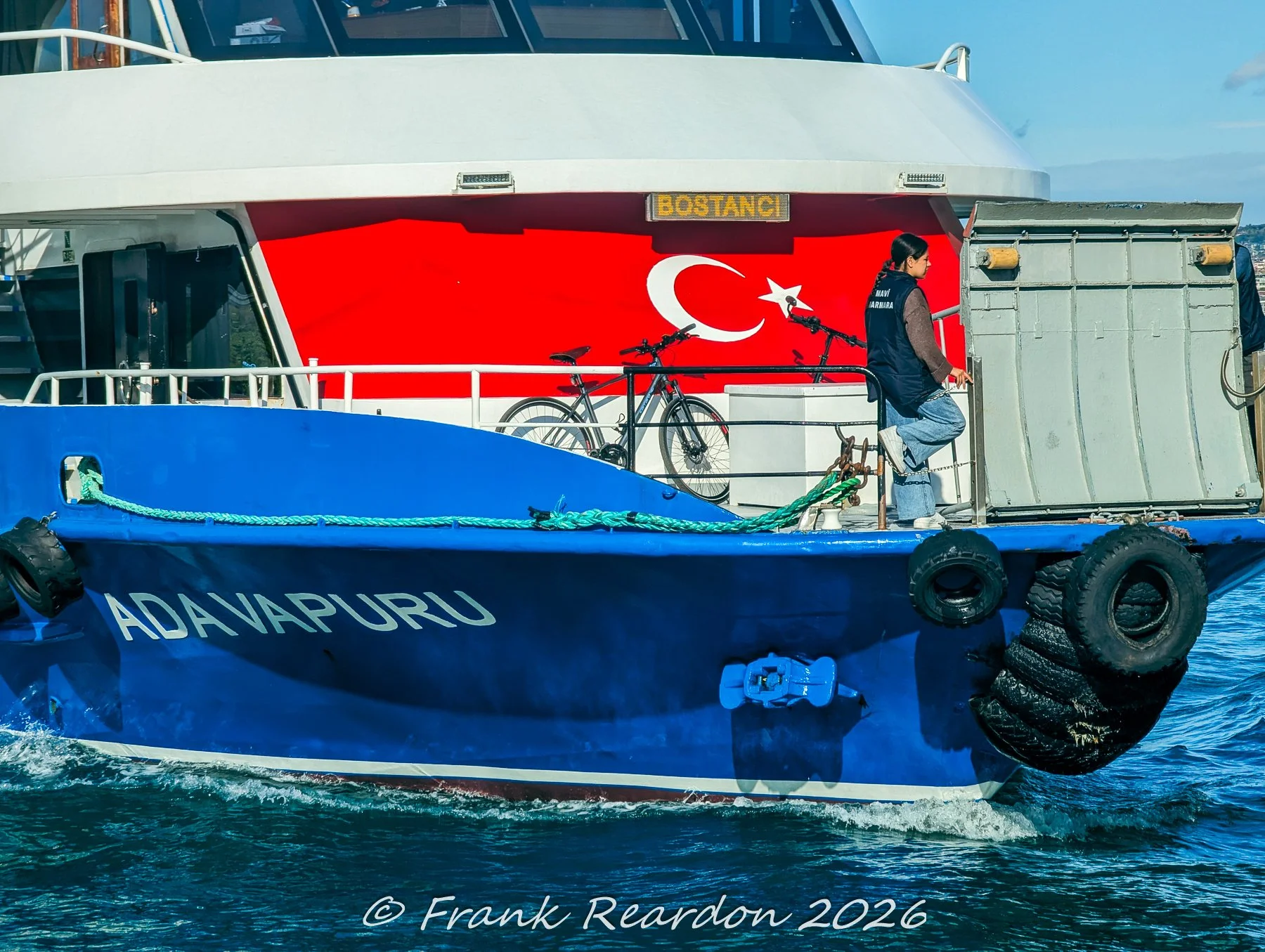 A frontal view of an Ada Vapuru passenger ferry showcasing a prominent Turkish flag painted on its hull alongside maritime ropes and safety tires.