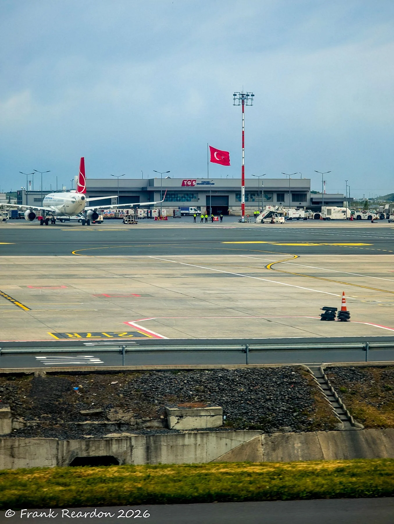 A bright daytime view across the expansive tarmac at Istanbul Airport, featuring an aircraft parked near the terminal building beneath the Turkish flag.