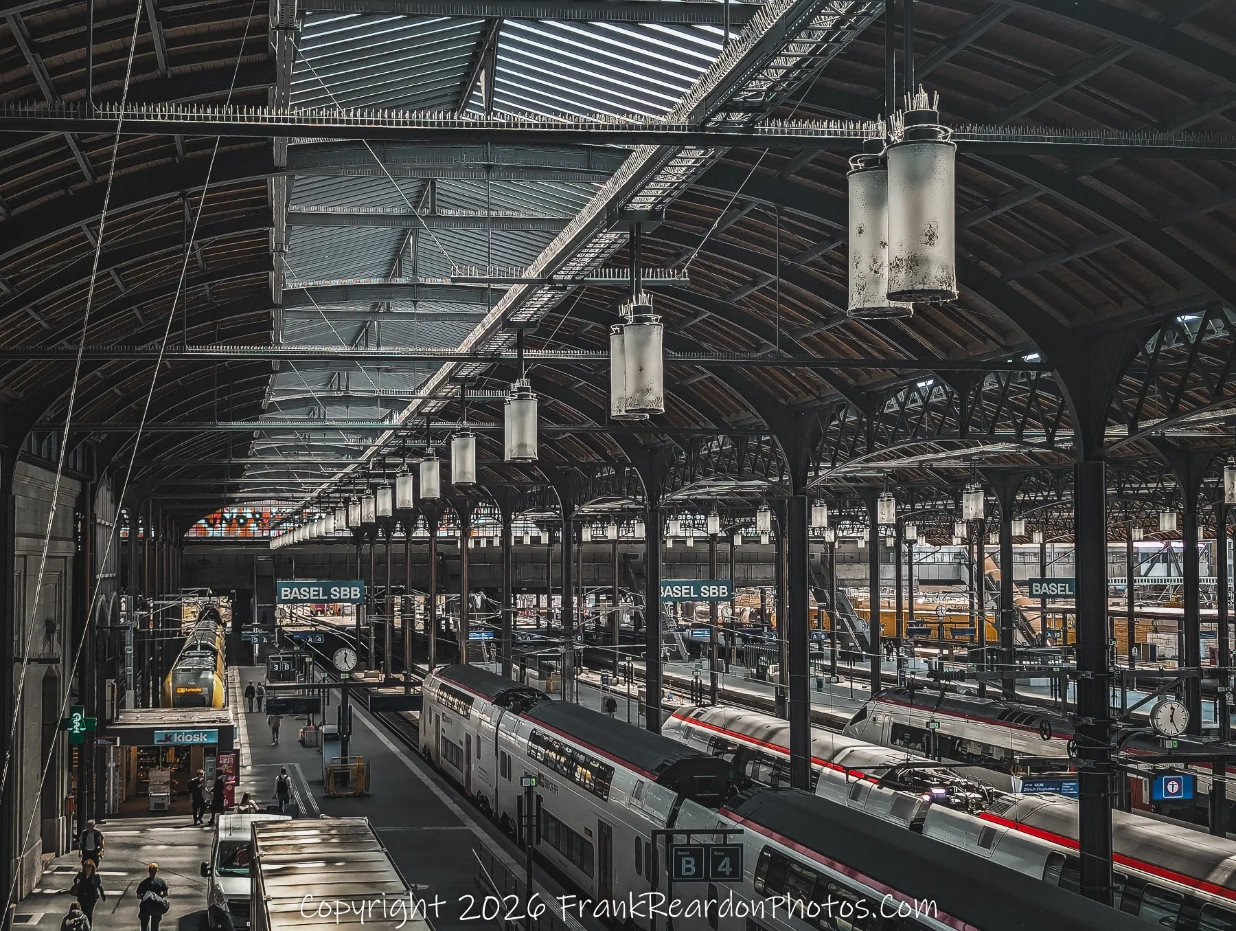 "The Great Hall" A wide-angle, high-perspective view of the Basel SBB train station, highlighting the intricate iron truss roof and rows of hanging industrial lights over the platforms.