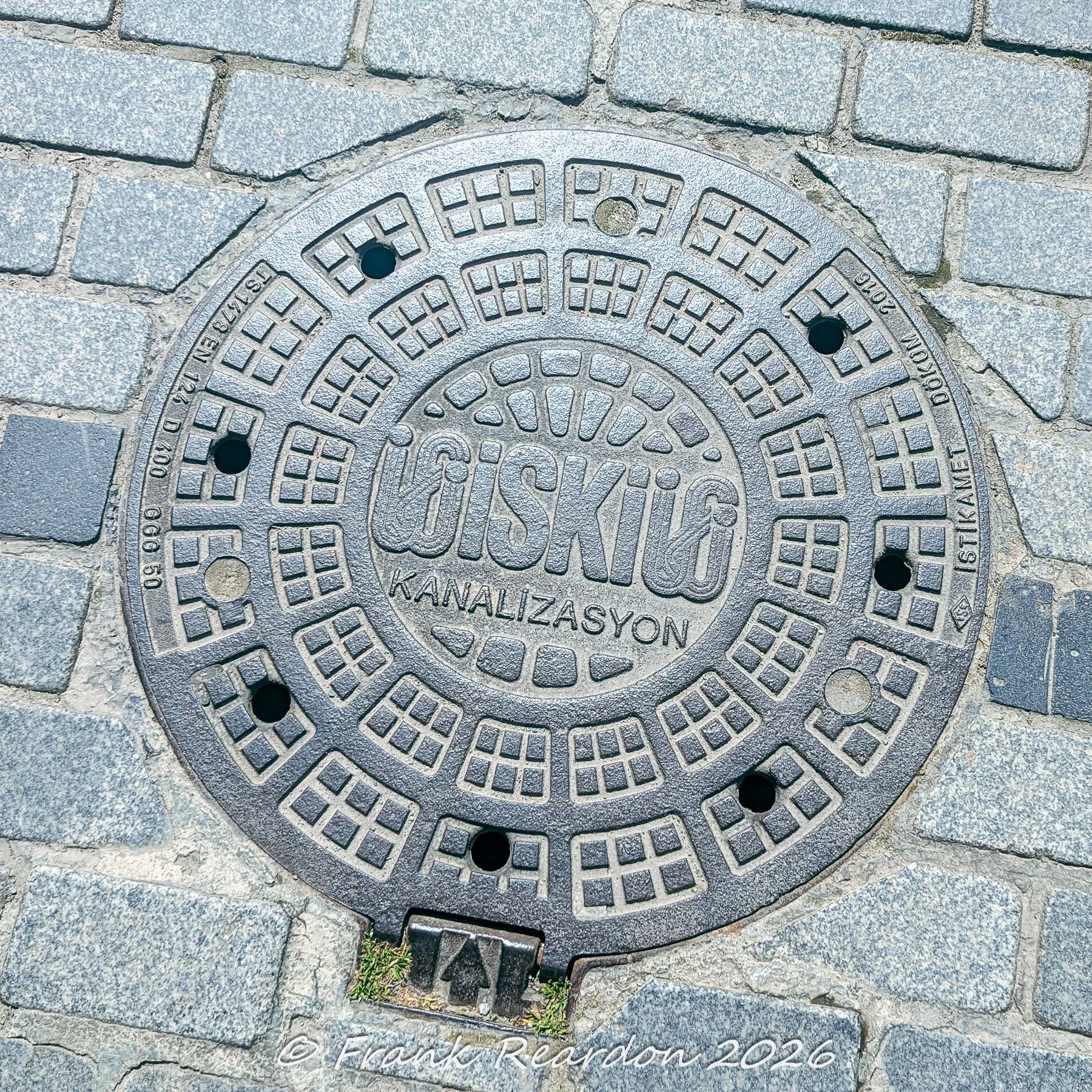 An intricate, heavy-duty iron manhole cover stamped with the Istanbul Water and Sewerage Administration (İSKİ) logo and canalization markings set into stone pavers.