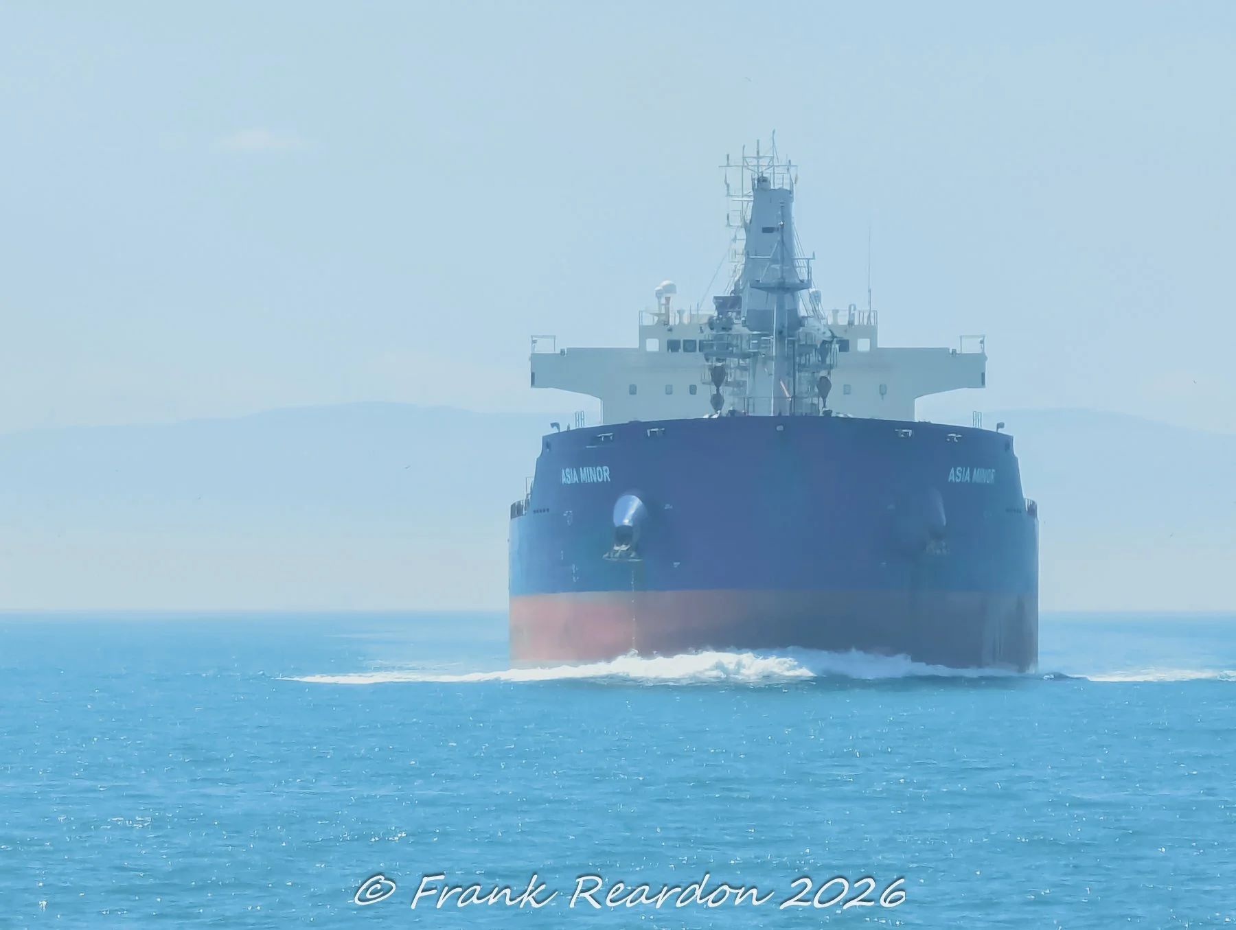 A forward view of the large cargo ship Asia Minor navigating through open coastal waters with a gentle white wake at its bow.