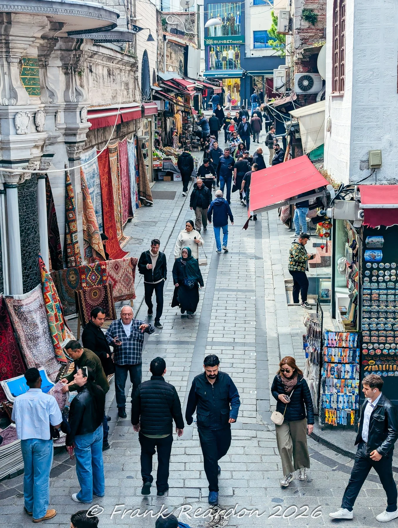 A busy sidewalk scene capturing shoppers, market displays, and traditional storefronts outside the Nuruosmaniye Mosque district.