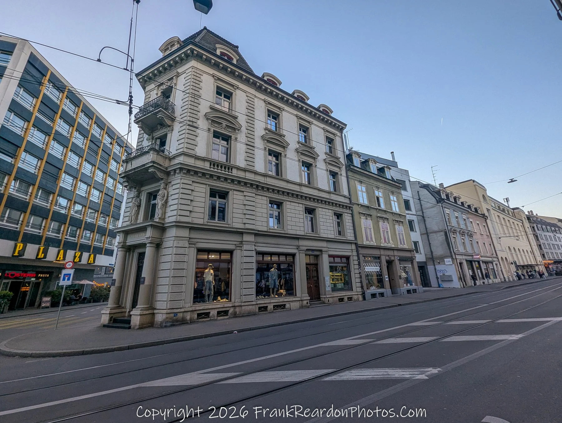 "Classical Urban Corner" An intersection showcasing historic multi-story buildings, including one with decorative stone statues on a balcony, under a clear sky.
