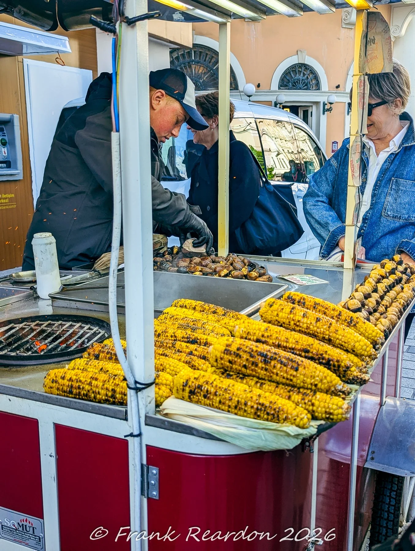 A street vendor grilling fresh ears of corn and roasting chestnuts on a traditional cart setup outdoors.