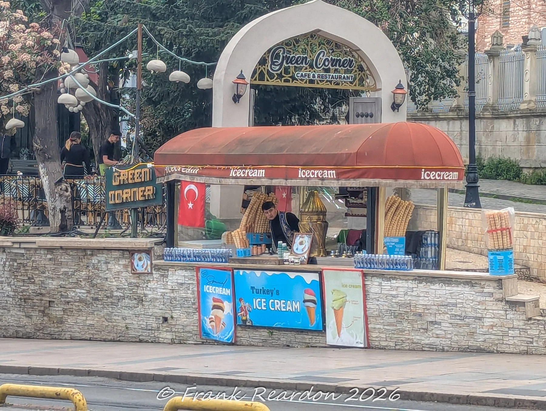 A vibrant street food cart selling traditional Turkish ice cream, positioned under a red awning near the Green Corner Cafe & Restaurant.