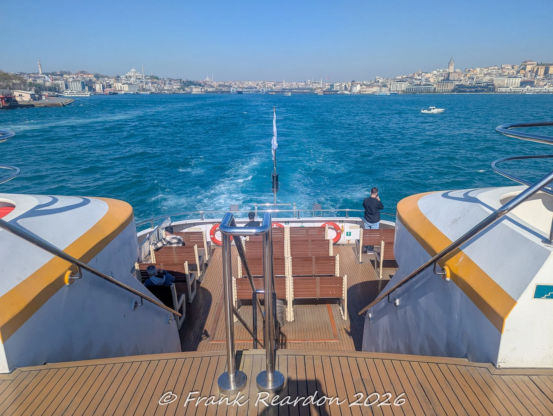 Looking backward from the stern of a moving passenger ferry, capturing the churning white wake, the Turkish flag trailing behind, and distant city views.