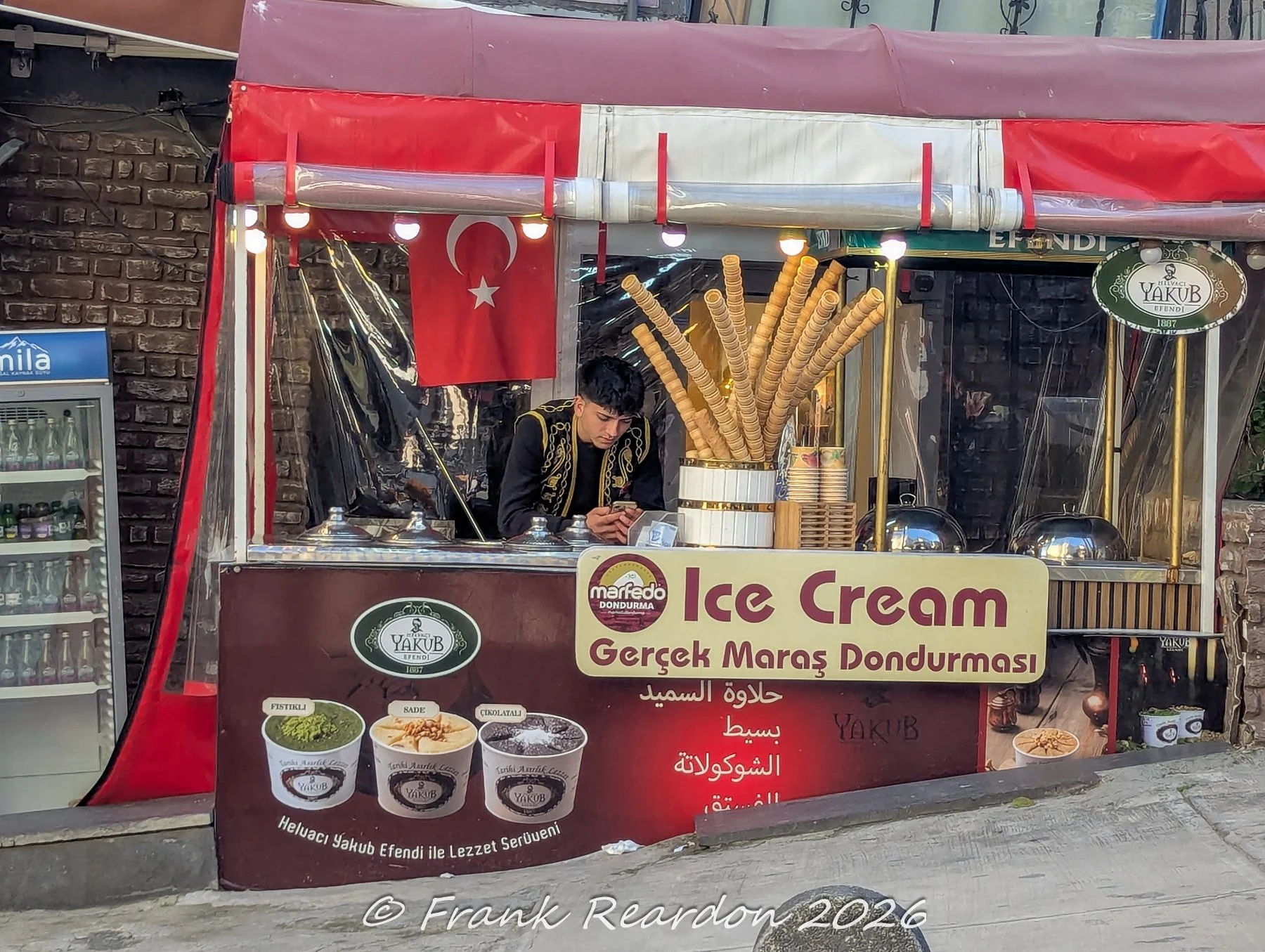 A street vendor operating a traditional Maras ice cream (dondurma) cart adorned with Turkish flags and a display of waffle cones.