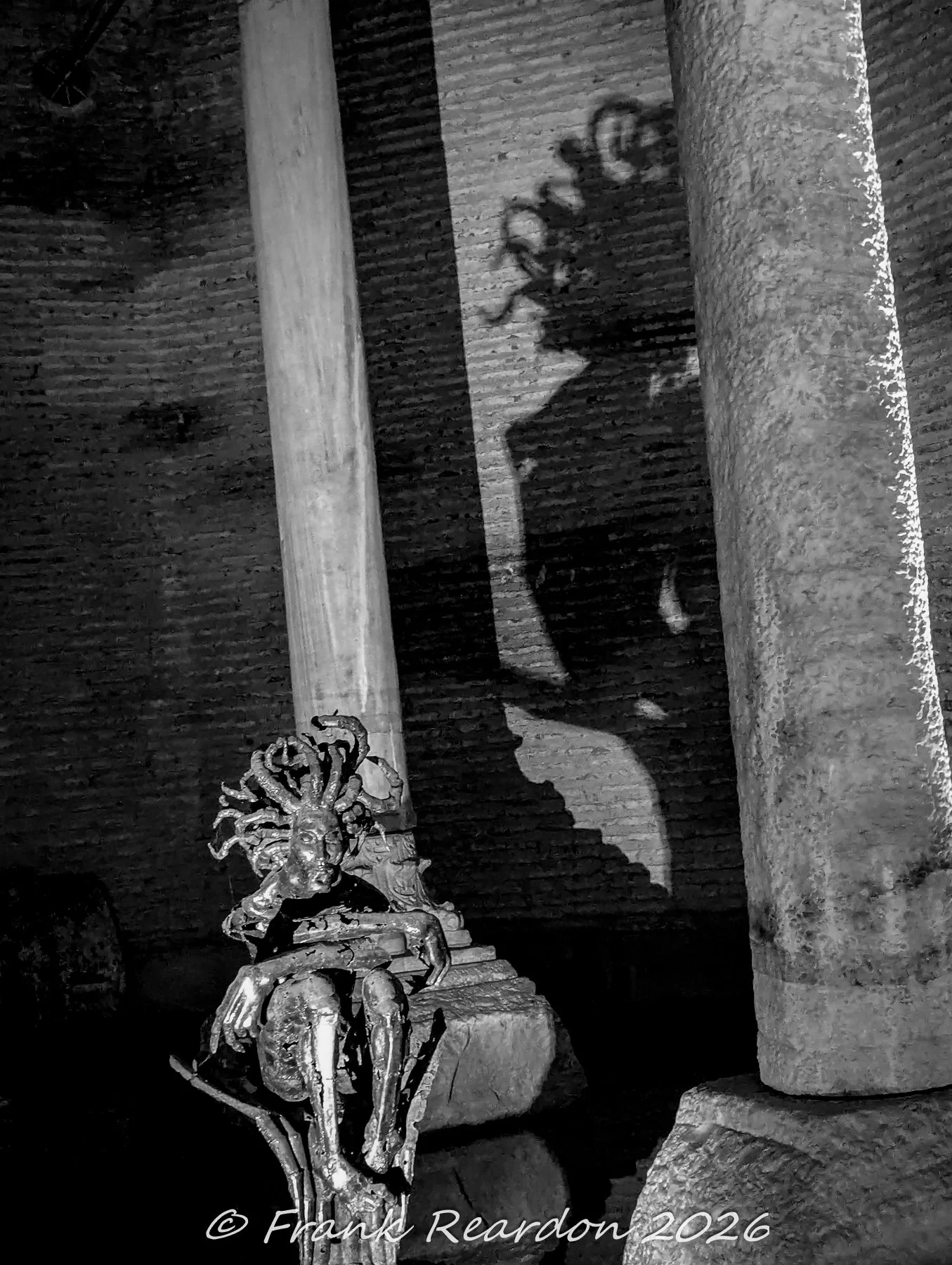 A black-and-white perspective showcasing the stylized Medusa art sculpture and the dramatic shadow cast against the ancient brick wall inside the Basilica Cistern