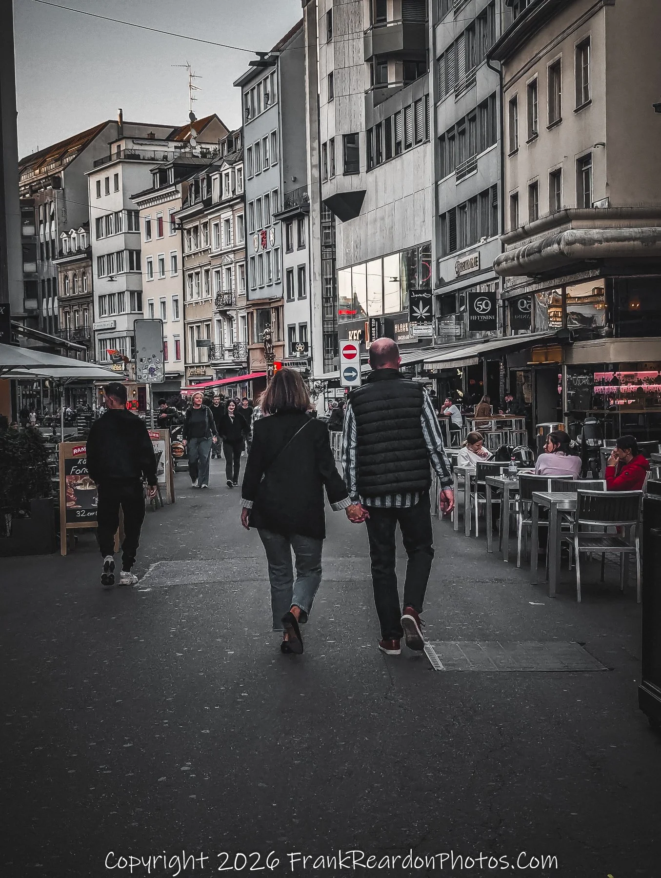 "Evening Walk in Basel" A couple holds hands while walking down a bustling city street lined with shops and outdoor restaurant seating, captured during the twilight hours.