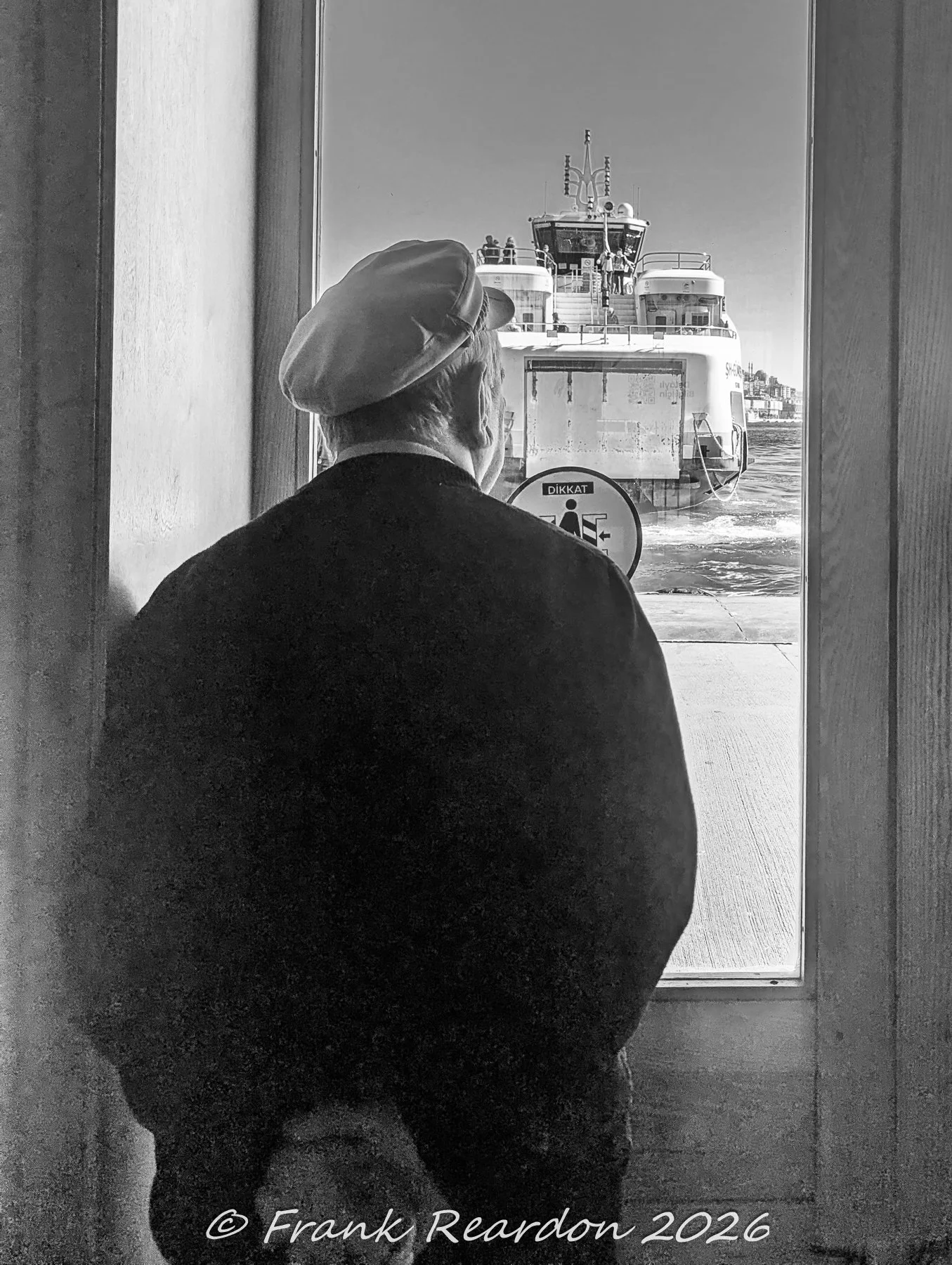 A black-and-white shot from inside a terminal or doorway of a person in a cap looking out at a incoming Bosphorus passenger ferry.
