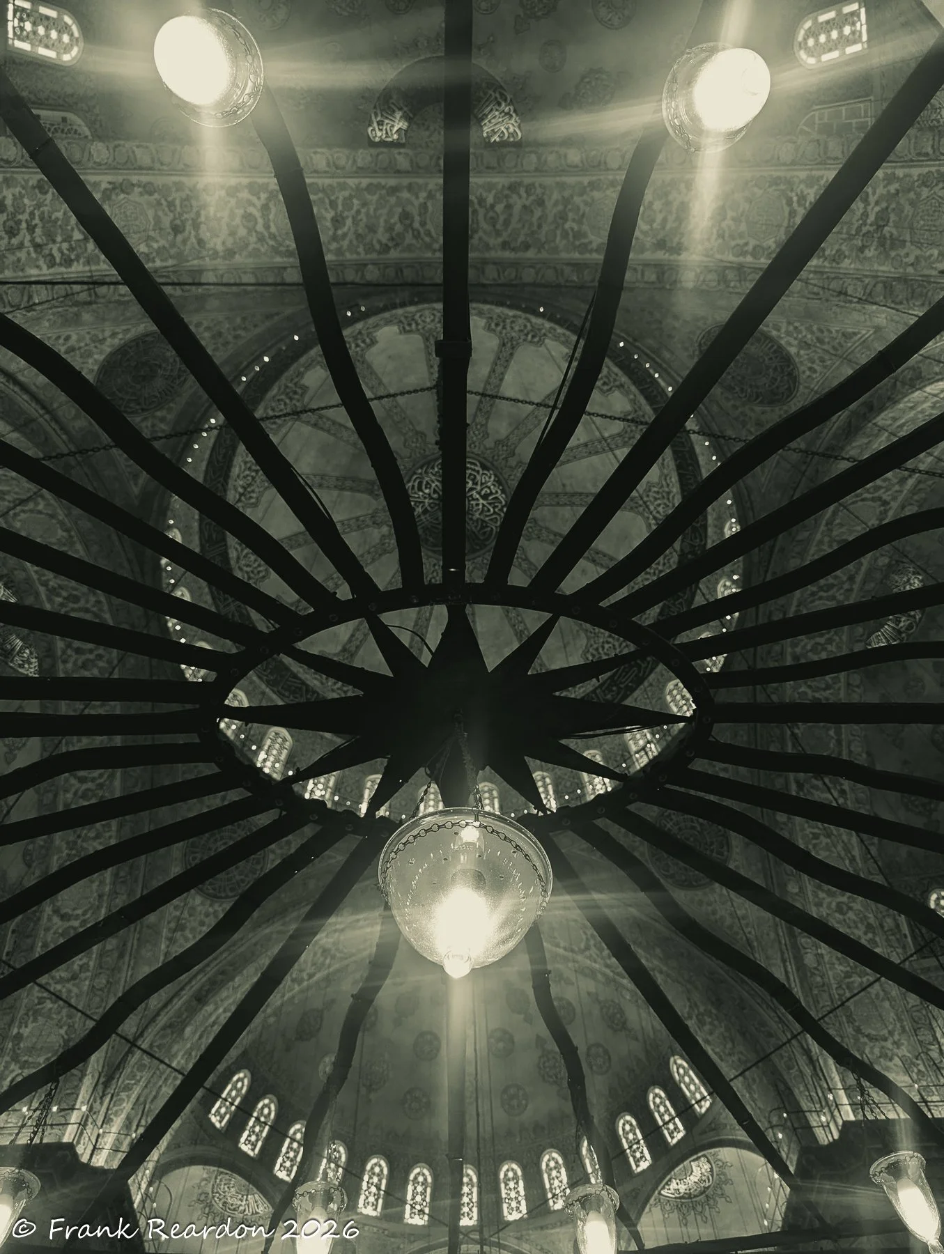 A striking upward look at the geometric metal frame of a large hanging light fixture radiating outwards beneath the mosque's upper windows.