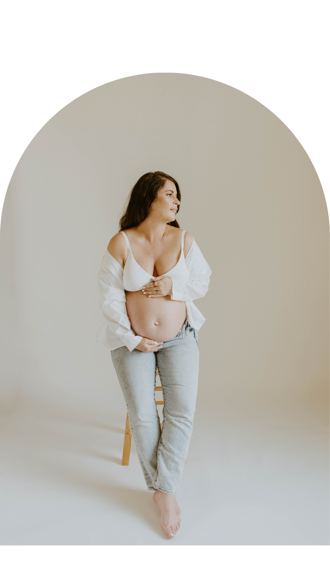 A pregnant woman seated on a wooden stool against a plain off-white background, wearing a white bra and light gray jeans, with a white shirt open at the shoulders. She is gazing to the side, with one hand on her belly and the other gently touching her chest.