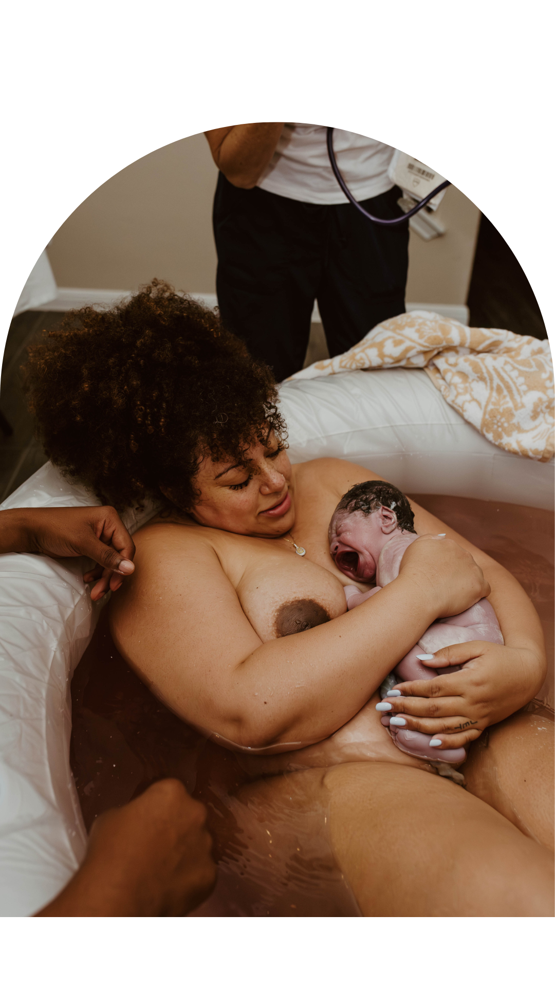 A woman with curly hair holds her newborn baby in a bathtub. The baby is crying. A person standing nearby is partially visible.