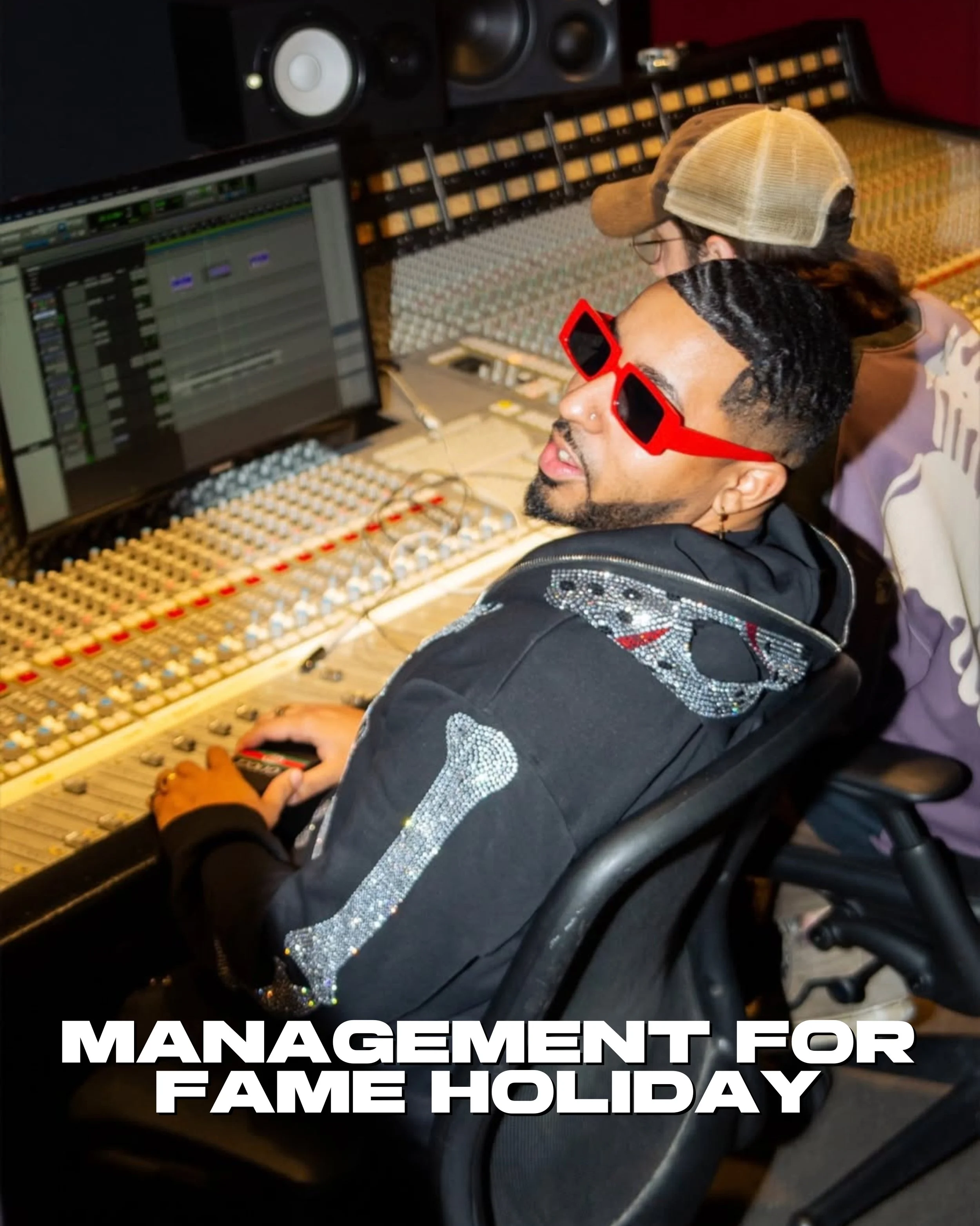 Fame Holiday with styled hair and red sunglasses working in a music recording studio, sitting in front of audio mixing equipment, with a woman in the background wearing a beige cap and glasses.