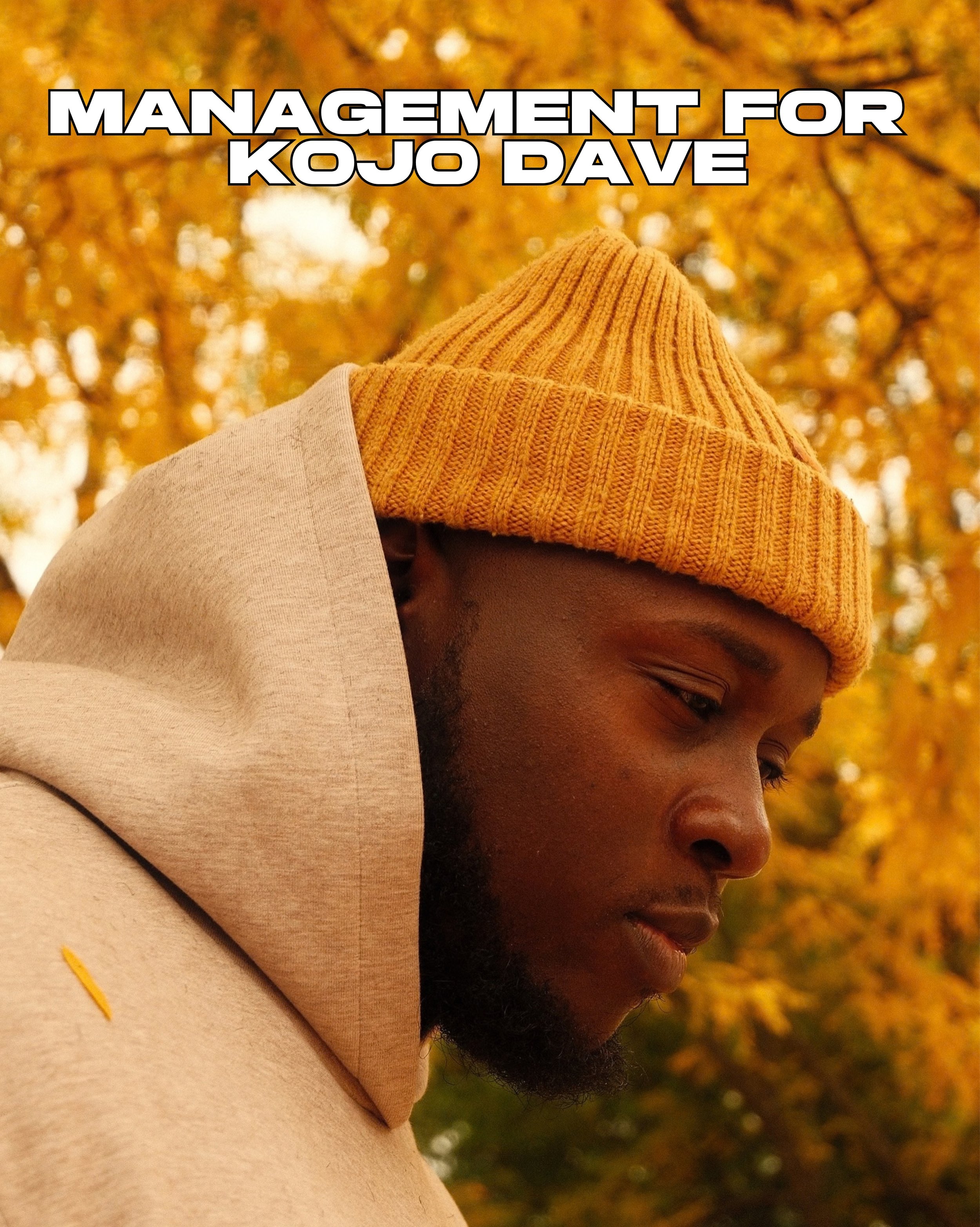 Close-up of Kojo Dave with a beard wearing a mustard yellow knit beanie and a beige hoodie, looking down with autumn trees in the background.