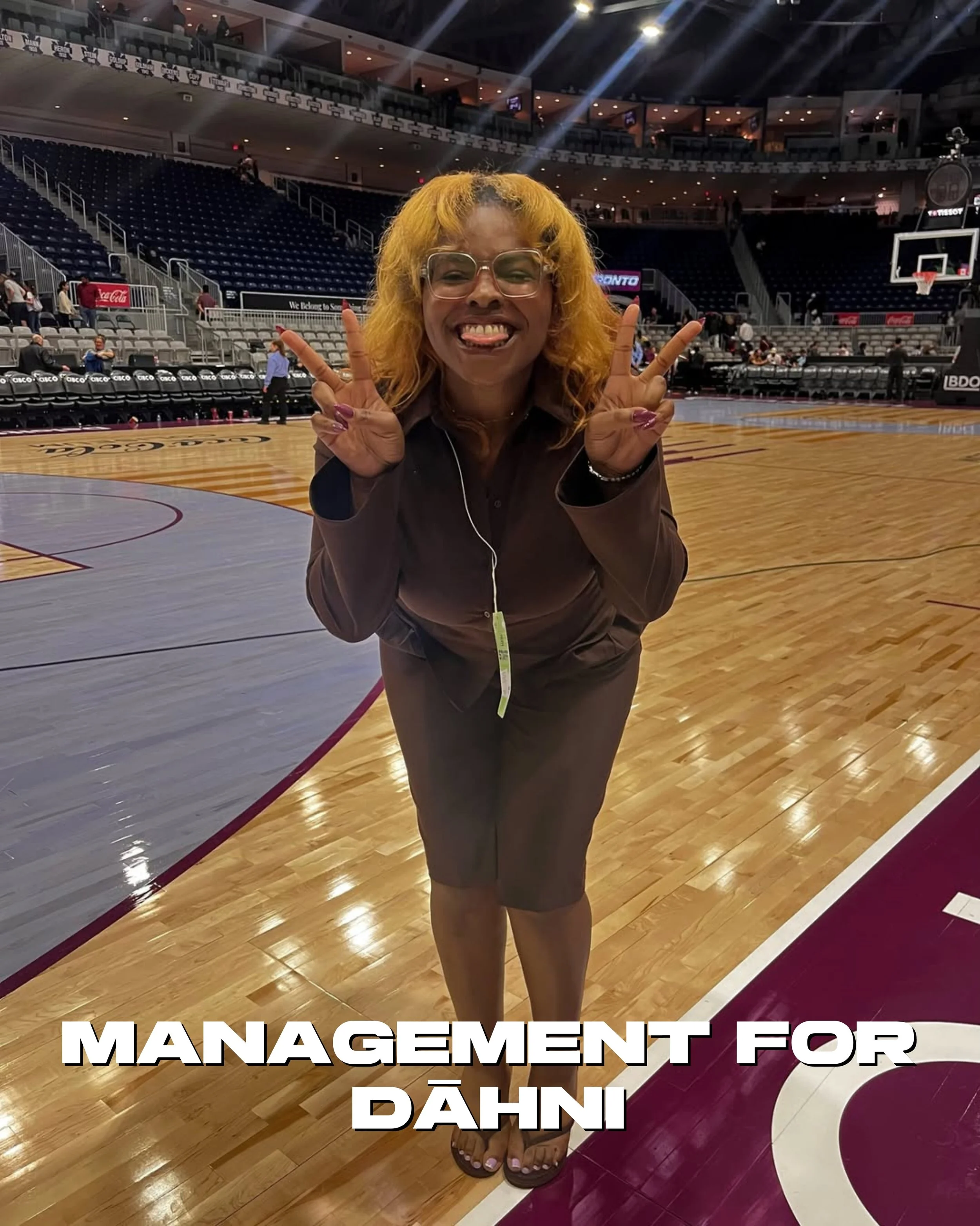 A woman with curly blonde hair, glasses, and a gray jacket making a peace sign with both hands, smiling and sticking out her tongue, standing on a basketball court with empty seats and some people in the background, and large white text at the bottom