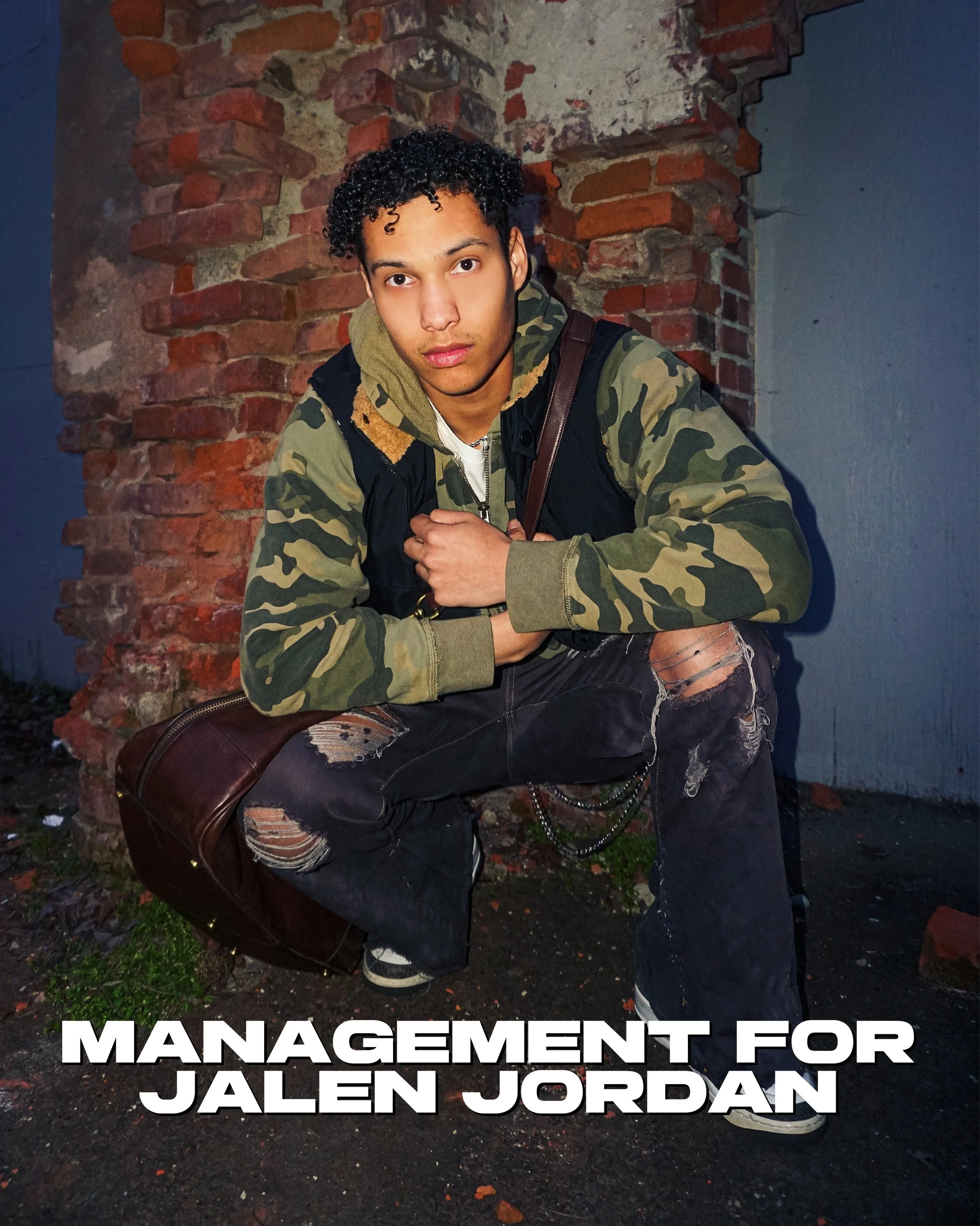 A young man with curly black hair wearing a camouflage jacket with a hoodie and a white t-shirt underneath, sitting on the ground against a brick wall. He has a brown leather bag next to him, and the ground is muddy with some green grass. The image a