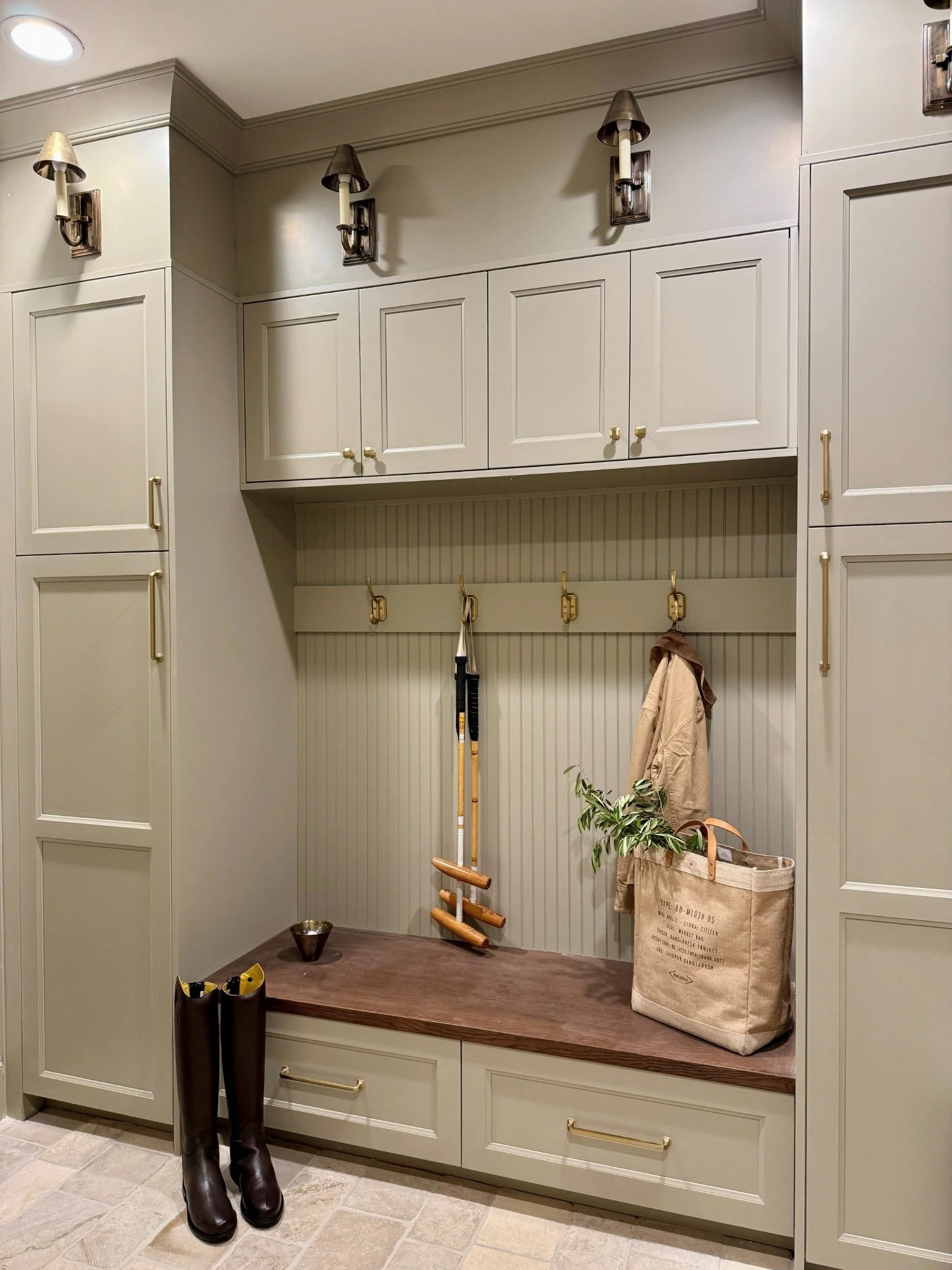 Mudroom bench styled with riding boots, croquet mallets, a hanging coat, and a canvas bag filled with greenery.