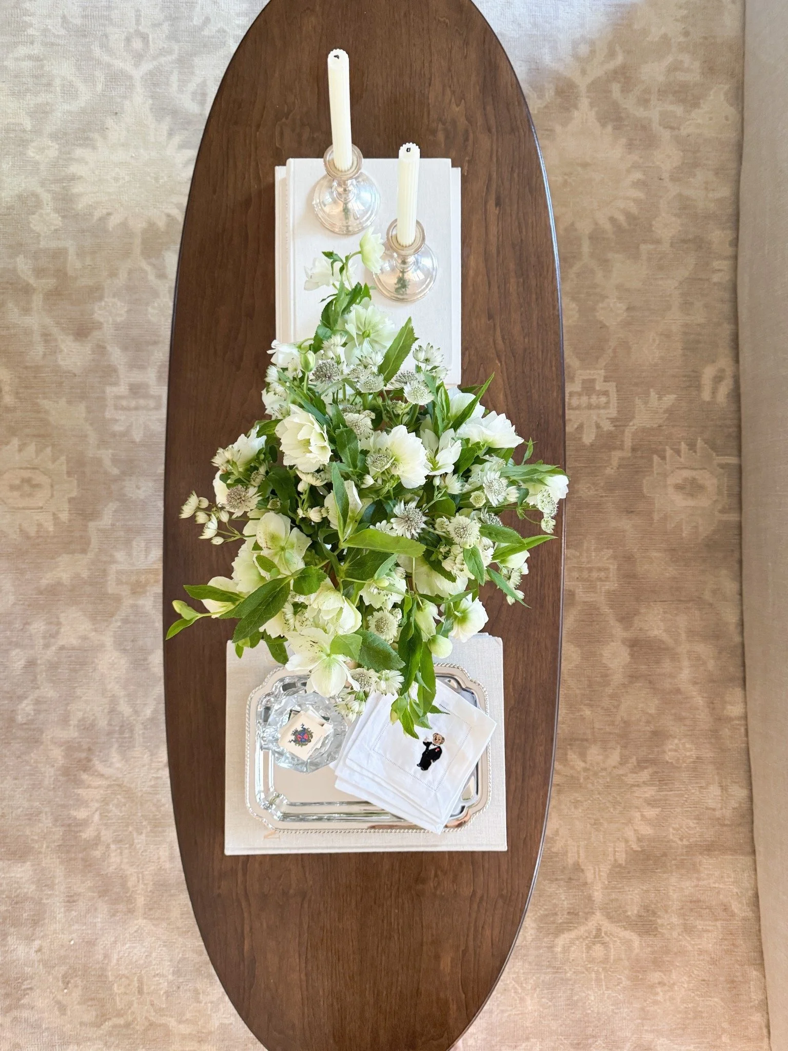 Overhead view of an oval wood coffee table with a white and green floral arrangement, books, and a tray.