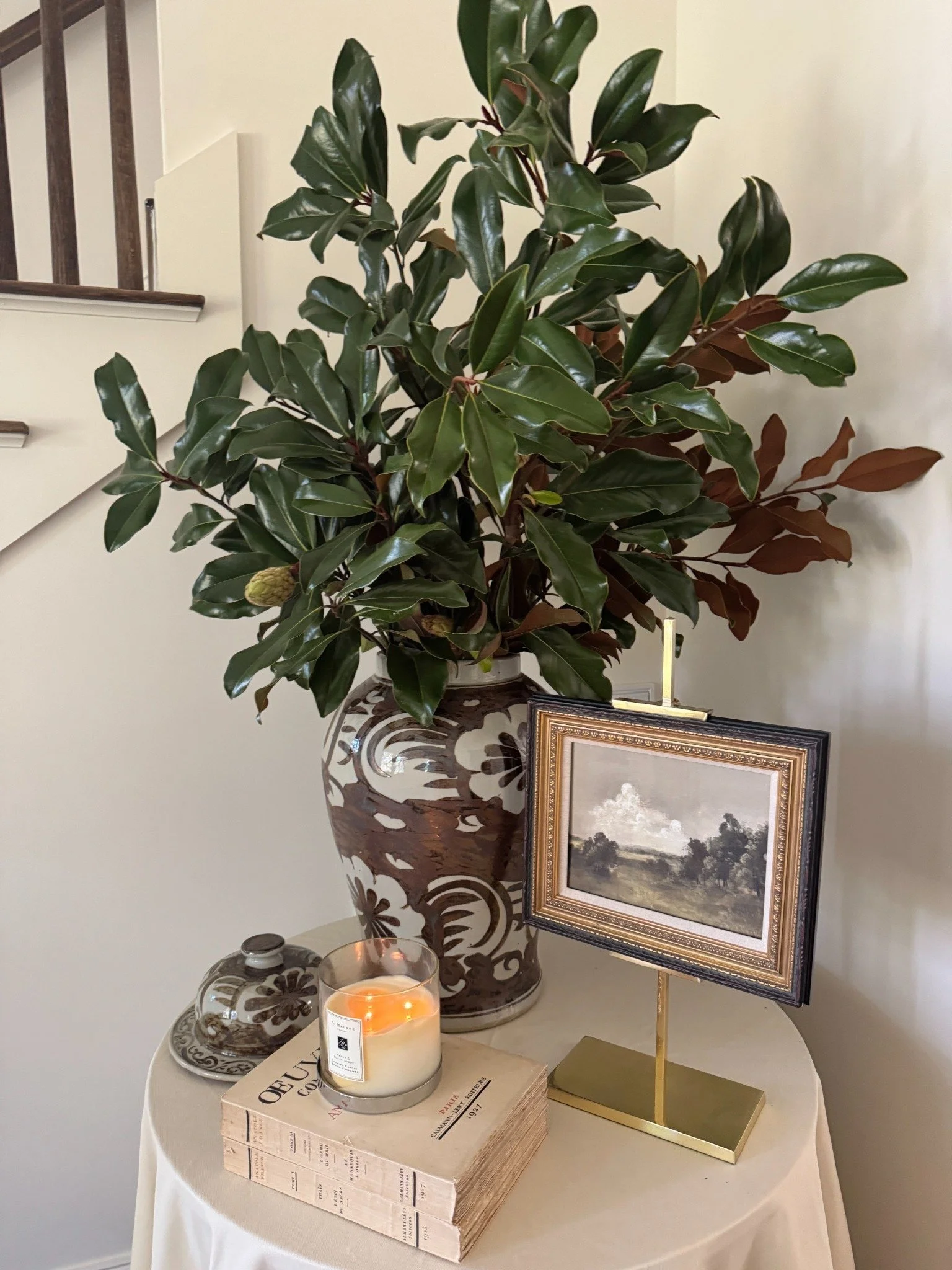 Magnolia branches in a patterned ceramic vase beside framed landscape artwork, stacked books, and a glowing candle.