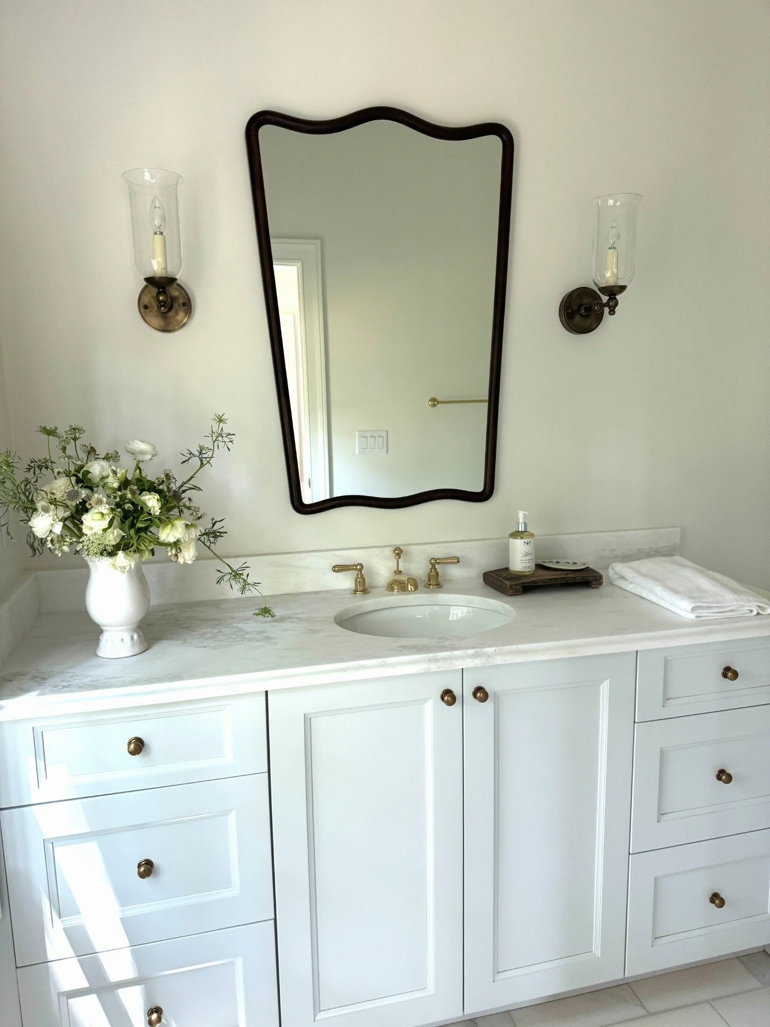 White floral arrangement beside a marble bathroom sink beneath a curved dark-framed mirror and glass hurricane sconces.