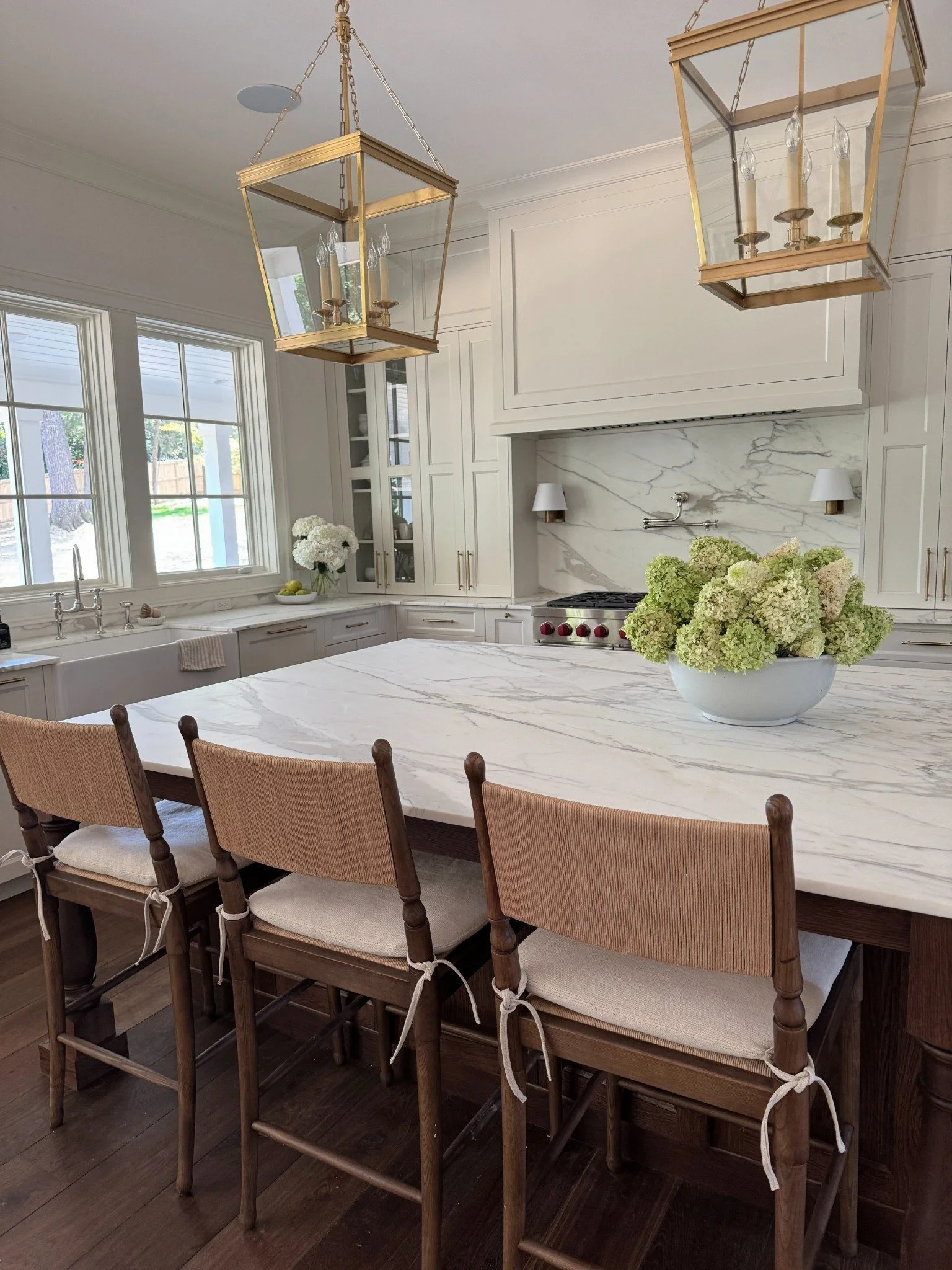White and green hydrangeas on a marble kitchen island beneath brass lantern pendants, with woven-back counter stools.