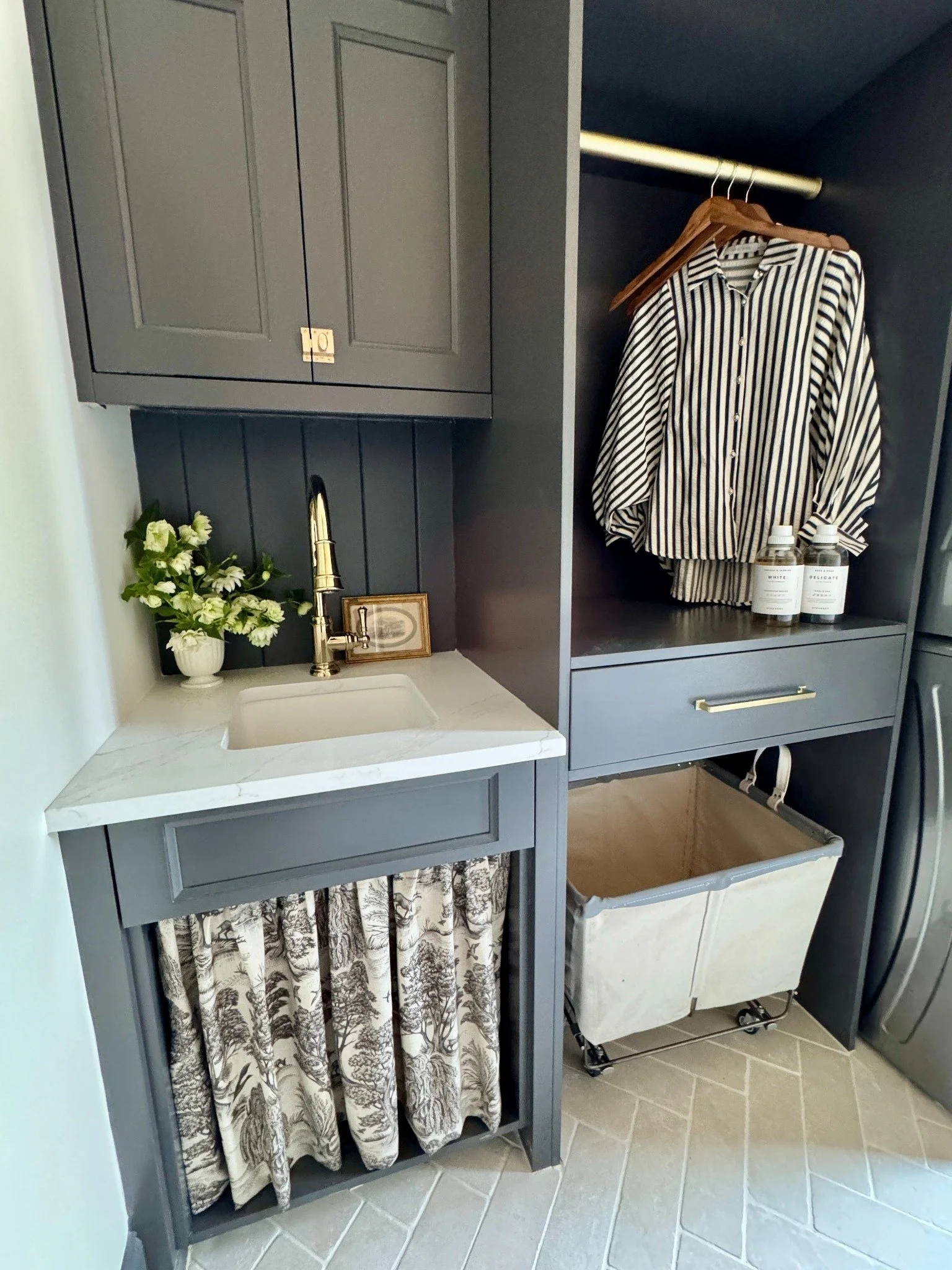 Dark-painted laundry cabinetry with a small floral arrangement, brass faucet, patterned sink skirt, and hanging striped shirt.