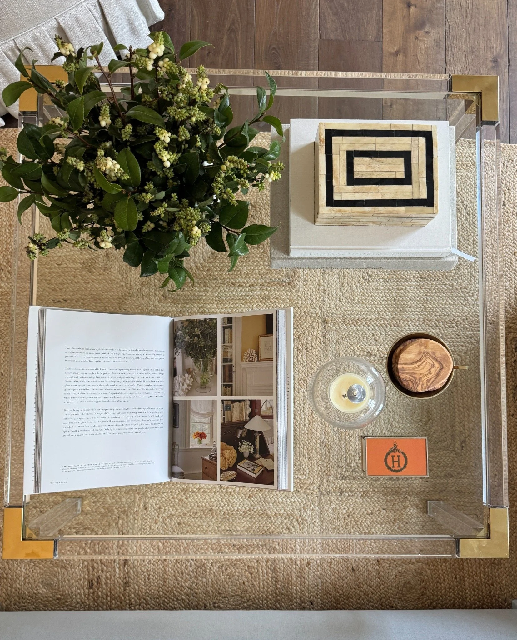 Overhead view of a glass coffee table with leafy branches, an open design book, a candle, and decorative boxes.