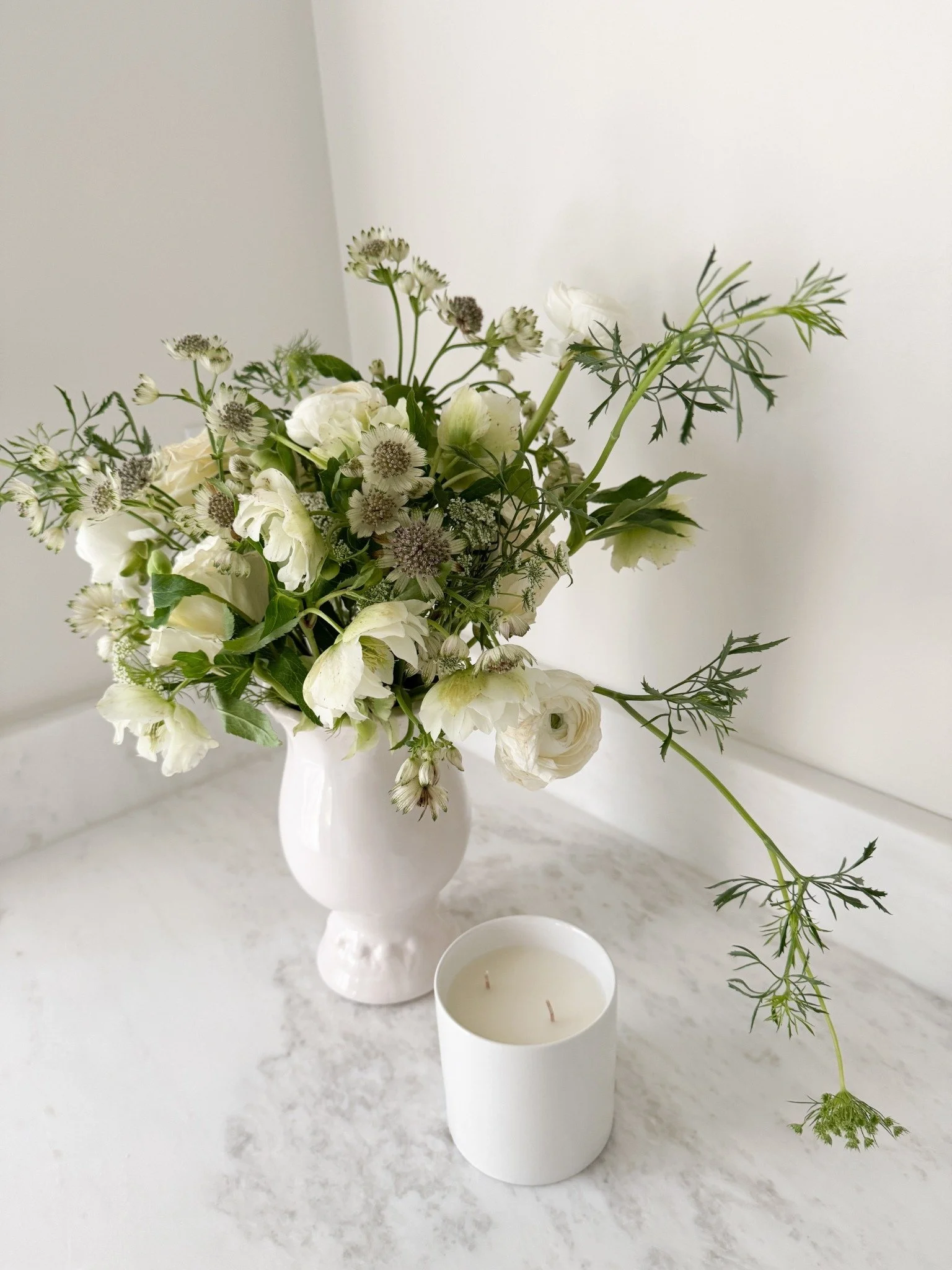 Loose white and green floral arrangement in a ceramic vase beside a candle on a marble surface.