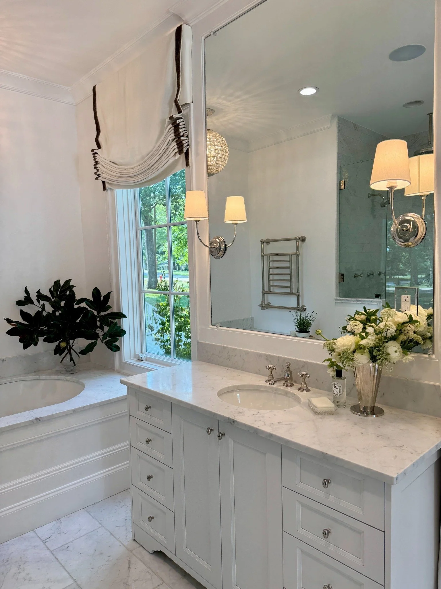 White floral arrangement in a silver vase beside a marble bathroom sink, with shaded sconces and a tailored Roman shade.