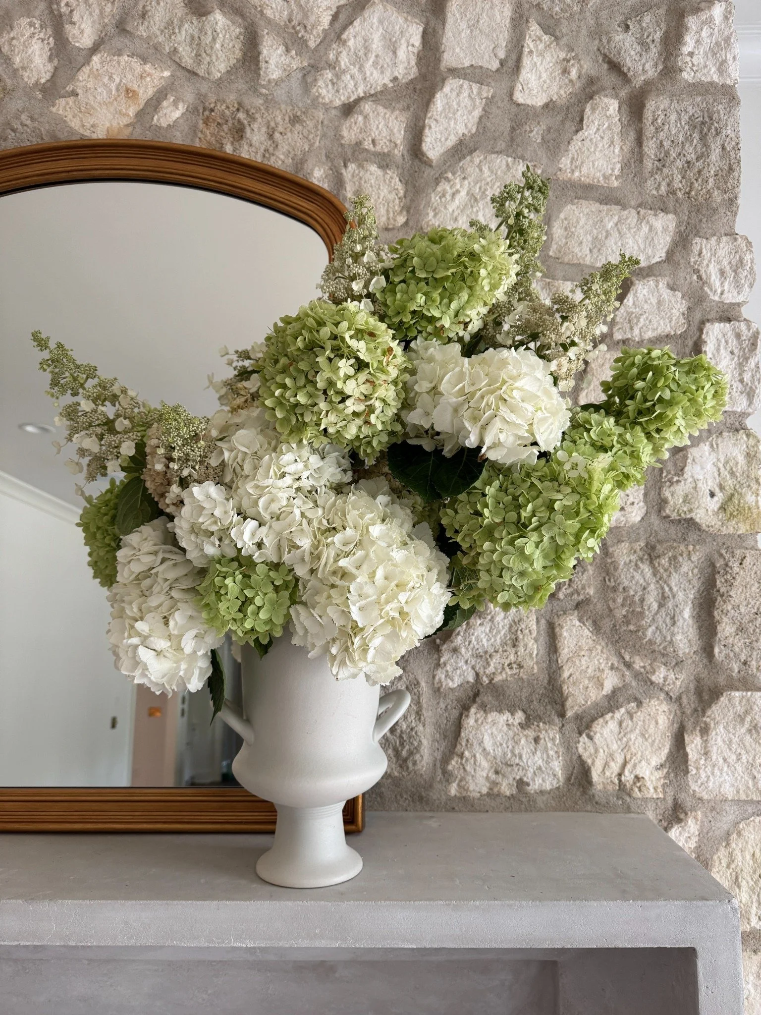 White and pale green hydrangeas with reaching flowering stems in a white urn on the stone fireplace mantel.