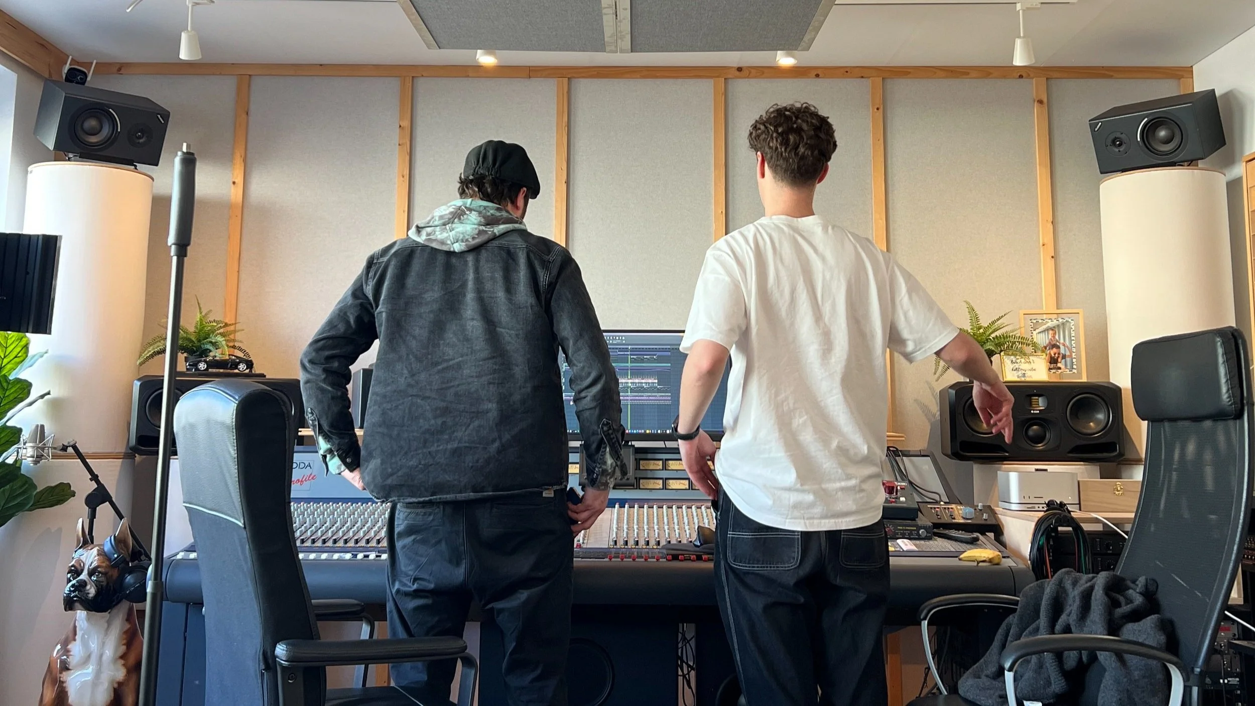 Two young men in a music studio working on audio production. One is wearing a black jacket and cap, the other a white t-shirt, standing in front of a mixing console with a computer screen showing audio editing software. The studio is equipped with large speakers, plants, and framed photos, with a dog sitting nearby.