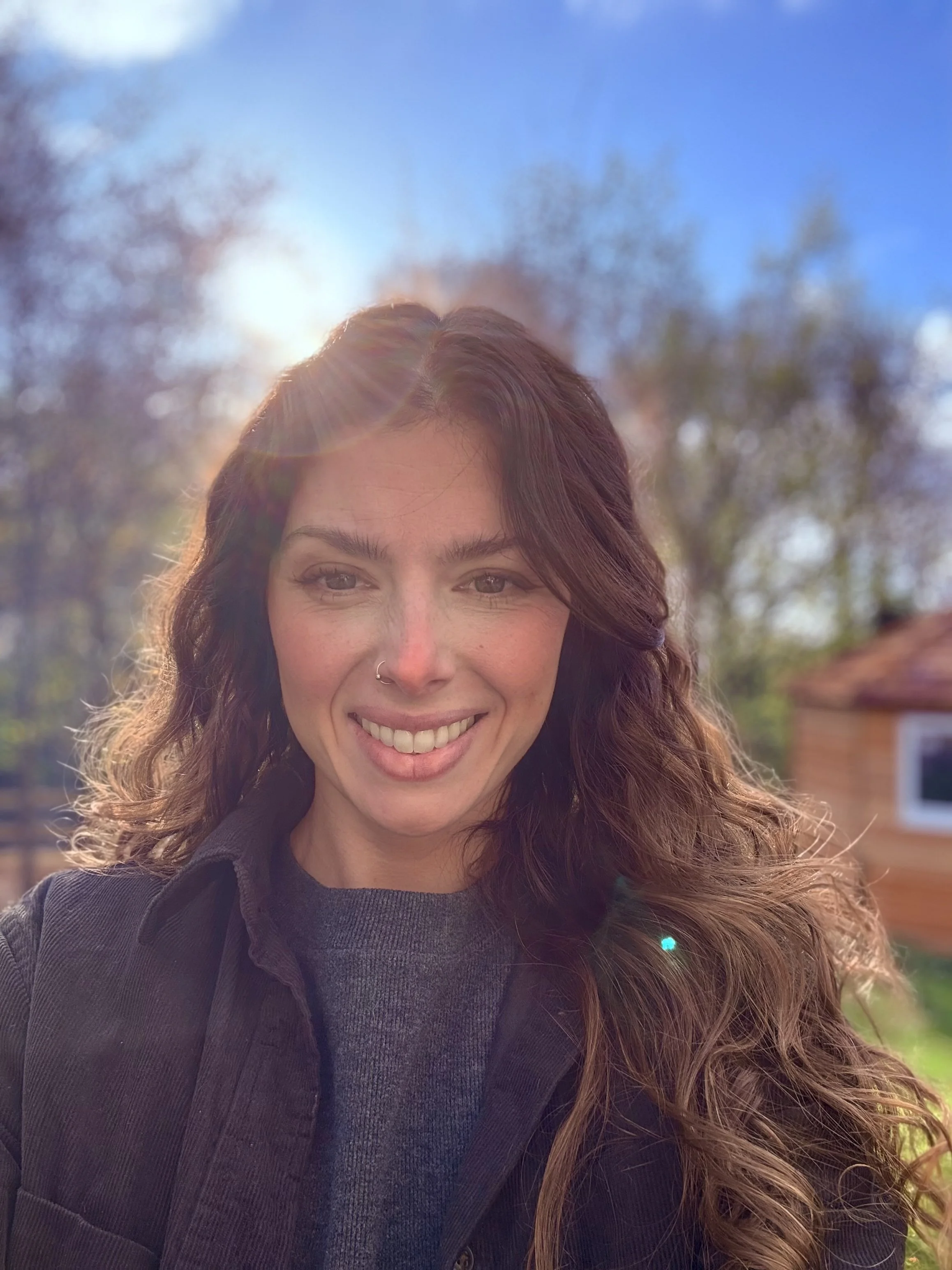 A woman with long, curly brown hair and a nose piercing smiling outdoors with sunlight, trees, and a house in the background.