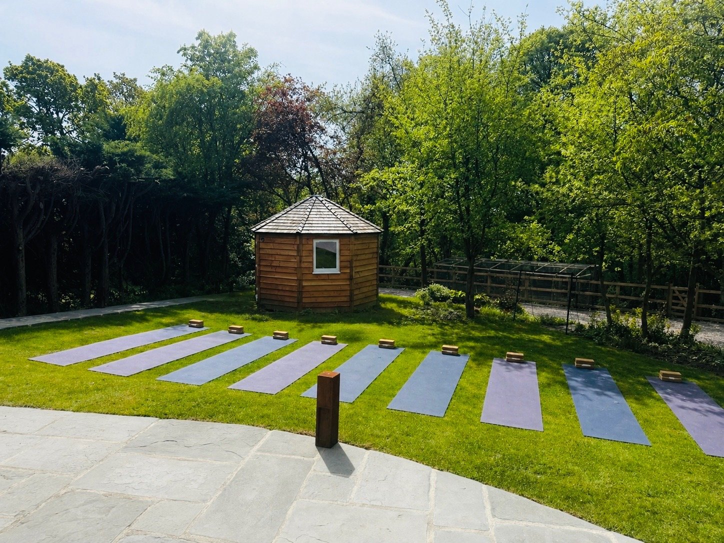 Outdoor garden space with seven yoga mats laid out on the grass, each with a brick block, surrounded by trees and a small wooden gazebo.