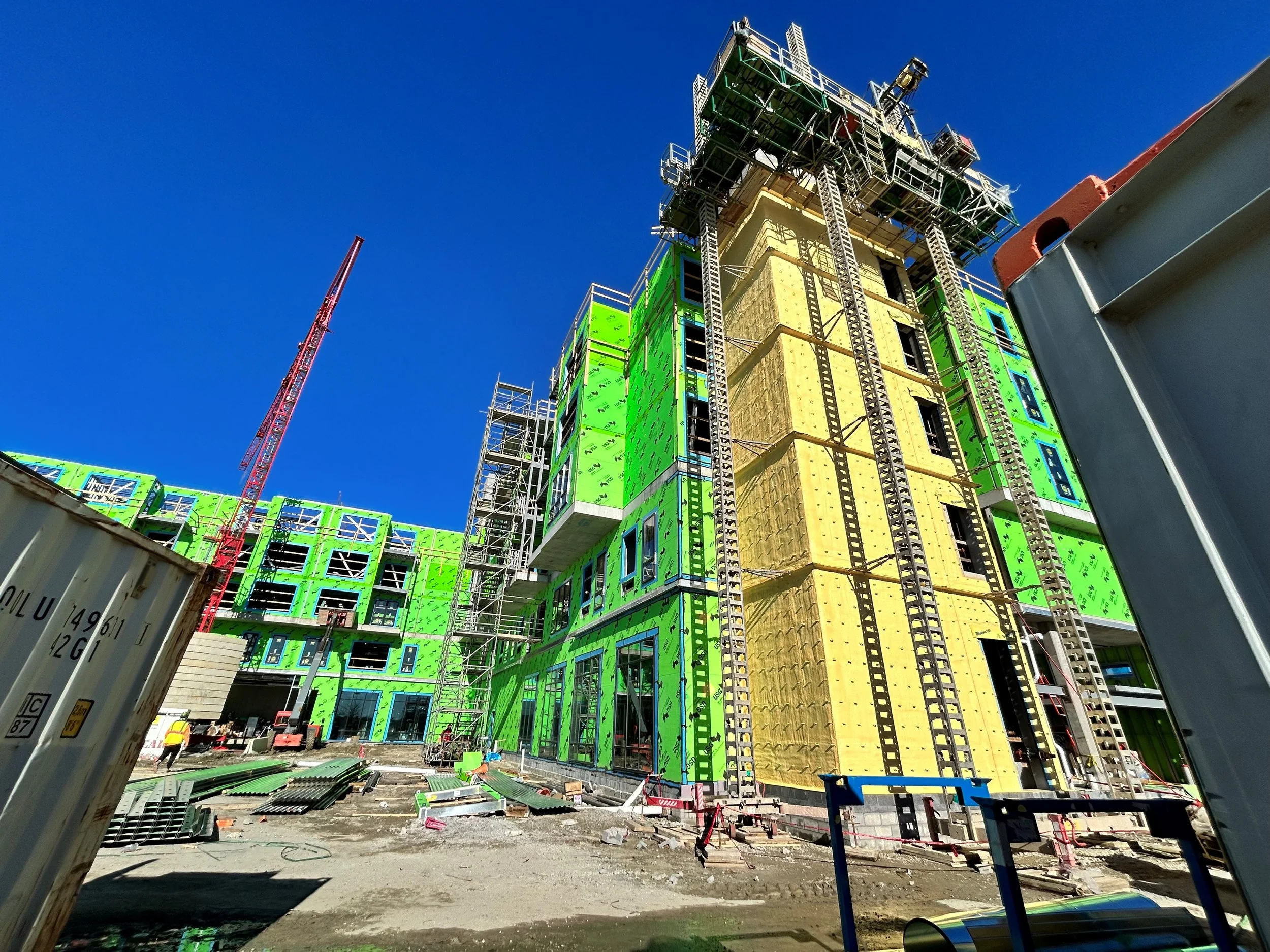 Construction site with partially built multi-story building covered in green sheathing, scaffolding, and workers, under a clear blue sky.