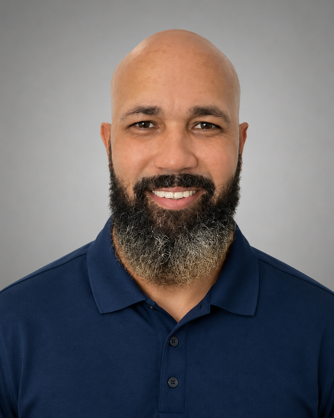 A man with a bald head and a full beard smiling, wearing a purple shirt and a yellow safety vest, outdoors on a bright day.